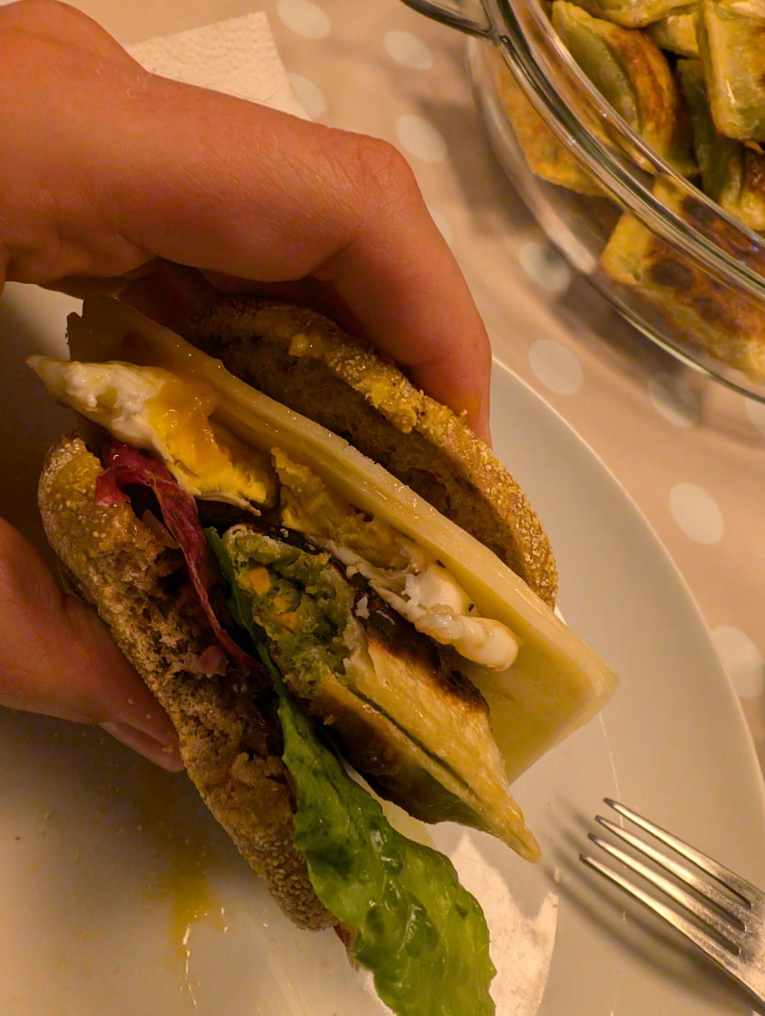 Auf dem Bild sieht man eine Hand, die einen Burger hält. Der Burger besteht aus verschiedenen Zutaten wie Salatblättern, gebratenem Ei, Käse und einer vegetarischen Maultasche. Der Tisch hat ein gepunktetes Tischtuch. Im Hintergrund ist eine Glasschüssel mit gebratenen Maultaschen zu sehen.