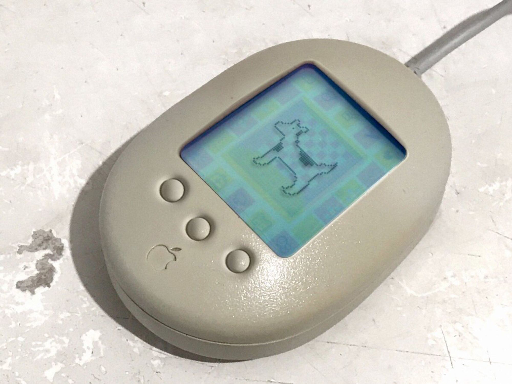 A picture of a mid 1990s bit of apple hardware in an oval shape with an adb cable out the back. On top of the device is a screen with a tamagotchi, except it’s Clarus the dogcow. Below the screen are three platinum beige buttons and an embedded (and slightly dirty) recessed apple logo.