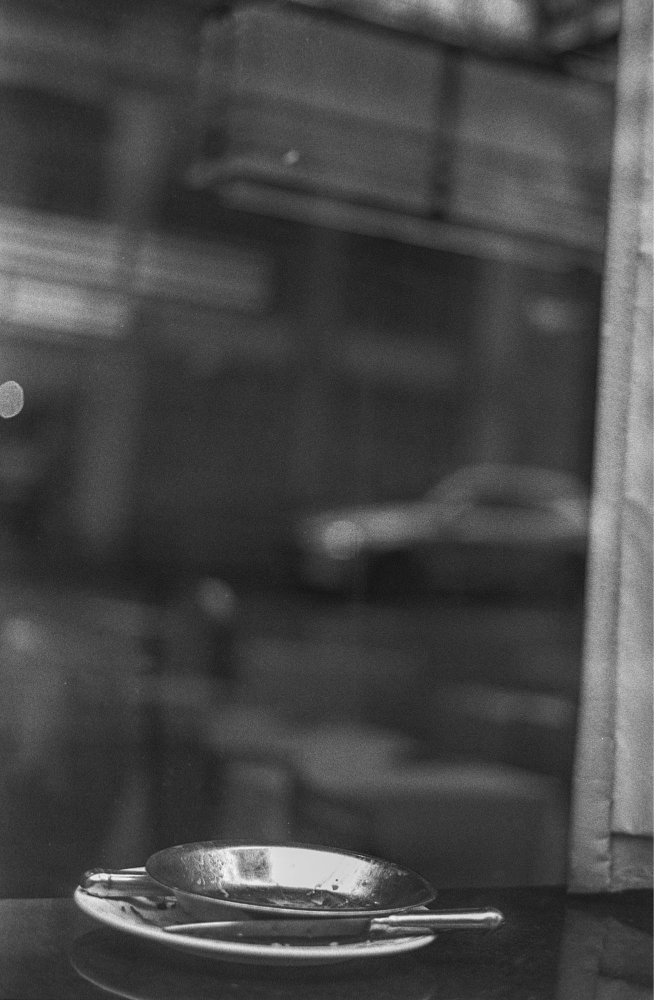 A black and white photograph of a pie pan with a knife laid across it behind a window in which a feathery bokeh of city chaos reflects the view: a dream of symphonic cacophony of the city; what the pie eaters may have scene.