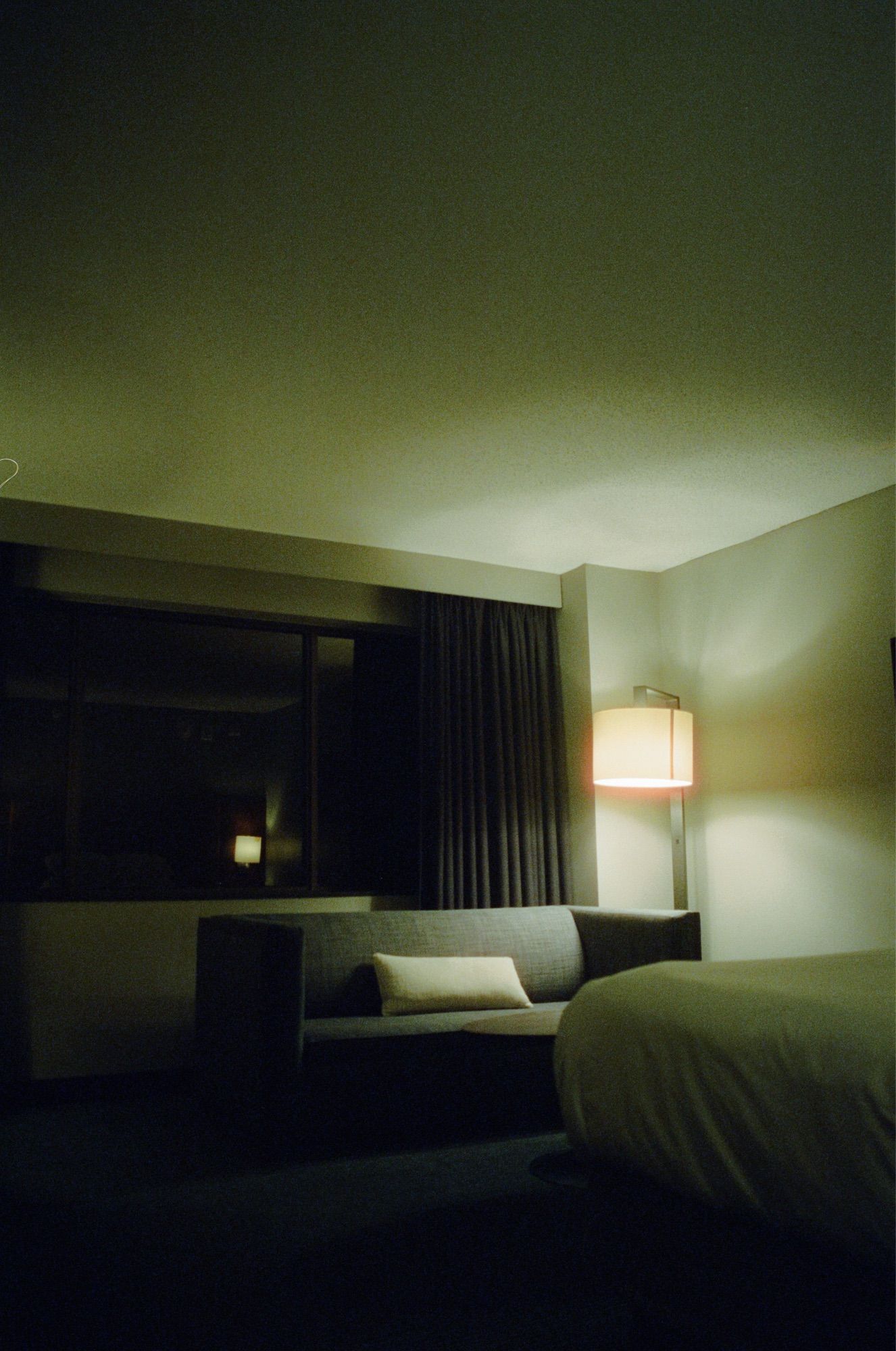 A color photograph of a couch facing a bed and watched over by the sizzling red halation glowing from a floor lap. Behind the couch is the darkened void of a major American city at night. The couch faces a bed.