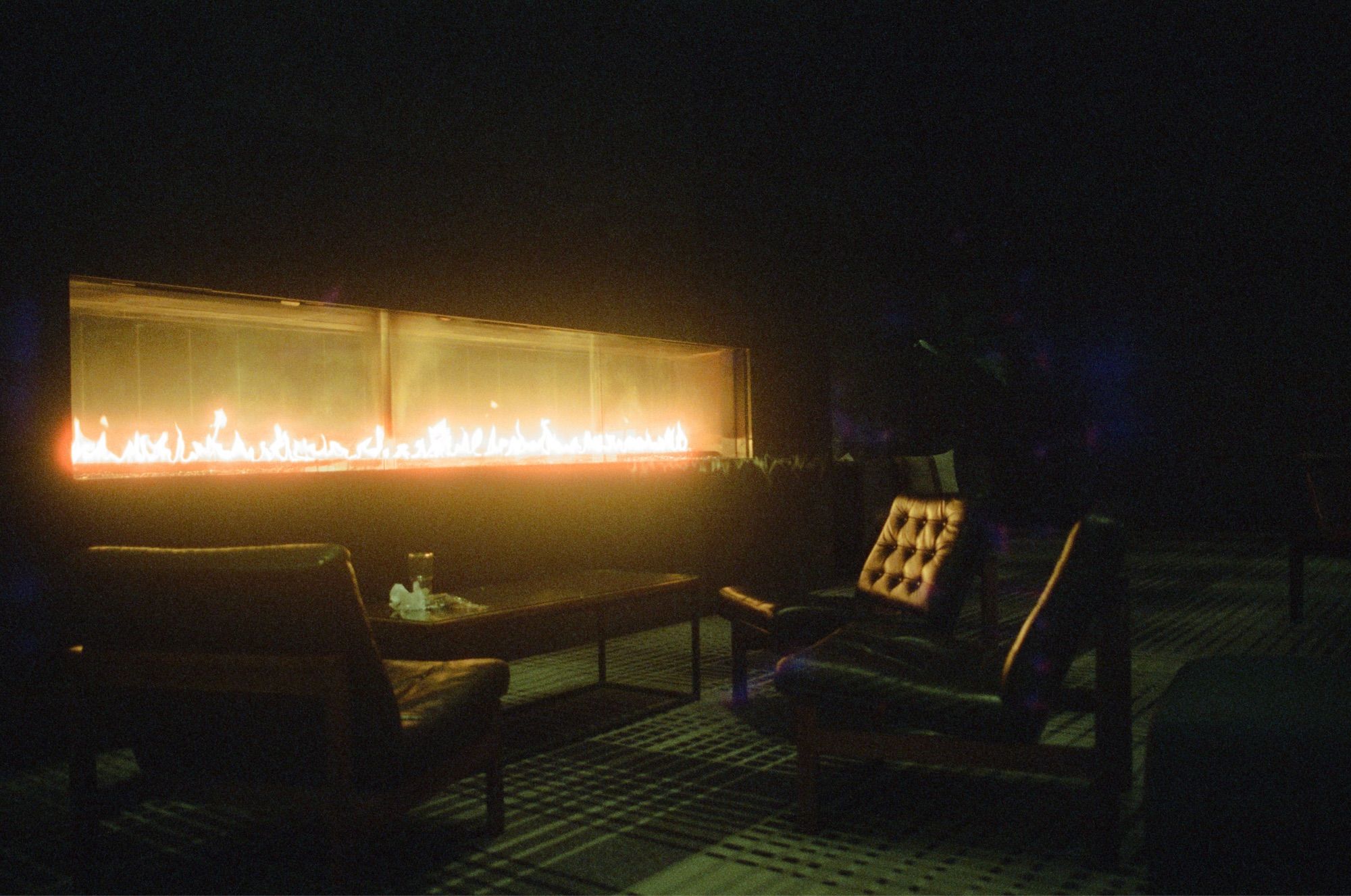 A color photograph of three Barcelona styled chairs sit around a long table before a length gas fireplace. An empty glass and some trash adorns the table: just moments ago people basked in the warmth and light here.