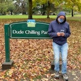 Stan in blue hoodie casually looking at his phone, leaning against a sign that reads "Dude Chilling Park." The ground is covered in yellow and orange autumn leaves.