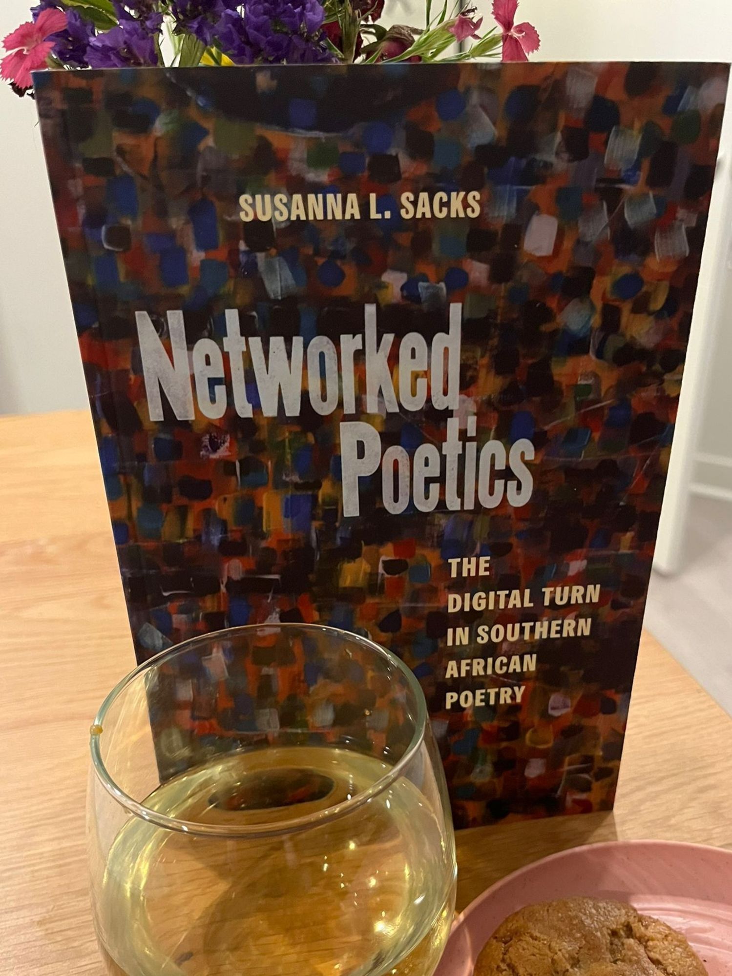 The book, "Networked Poetics," is pictured with a glass of wine, a plate of cookies, and some flowers