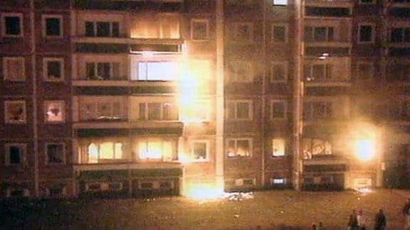 Ein Bild vom sogenannten Sonnenblumenhaus in Rostock-Lichtenhagen, dass durch Brandsätze Feuer gefangen hat. An der Fassade sind Flammen zu sehen, vor dem Haus stehen Menschen.