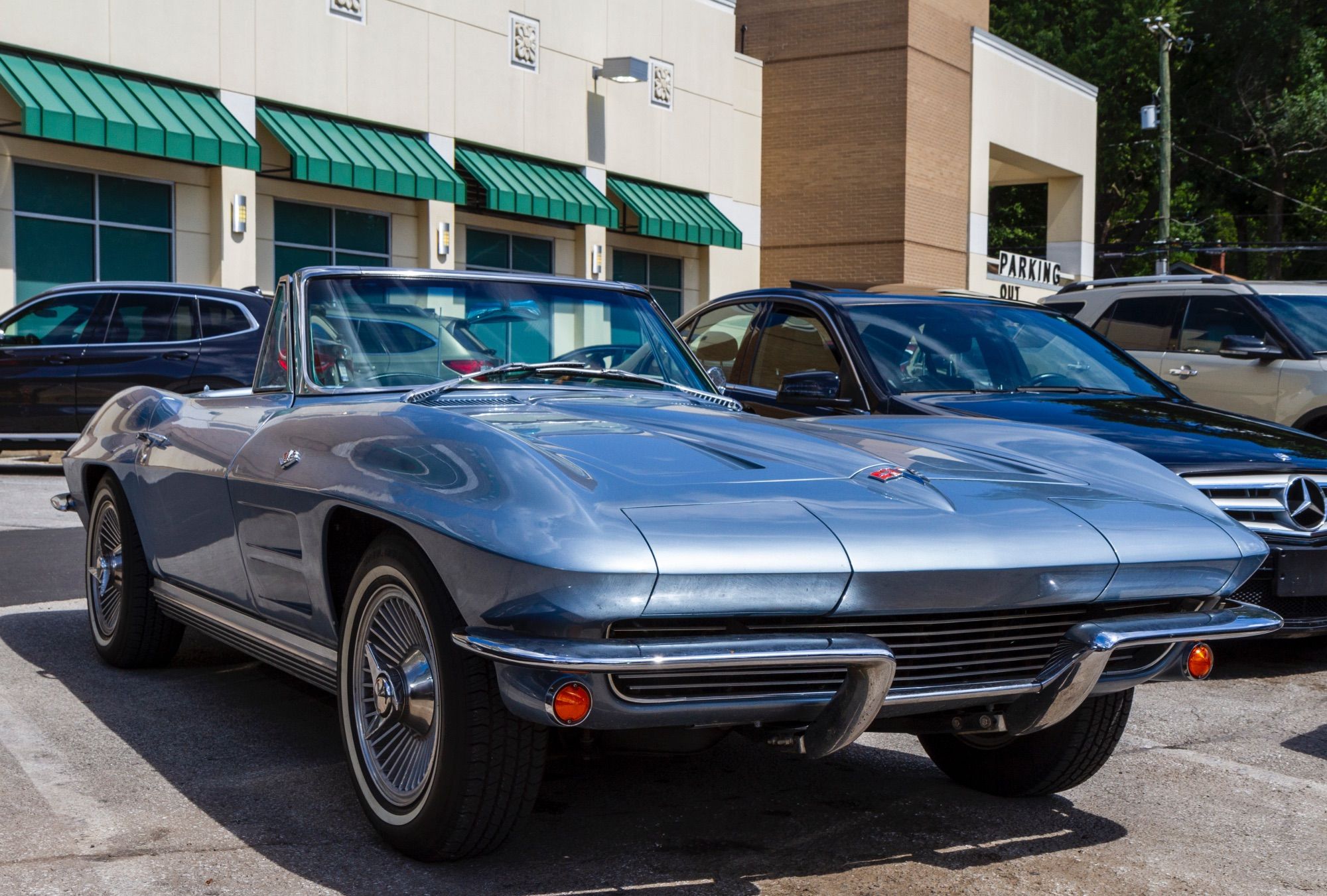 A low-angle view of a light metallic blue 1964 Corvette convertible with the top lowered, sitting in a Fresh Market grocery store parking lot.