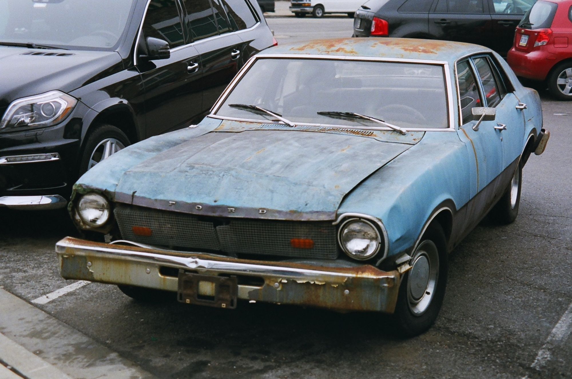 A view of an old Ford Maverick from the front left quarter. The grille has been replaced by wire mesh. The car is light blue and rusted in many places and all the sheet metal save the roof is slightly rumpled.