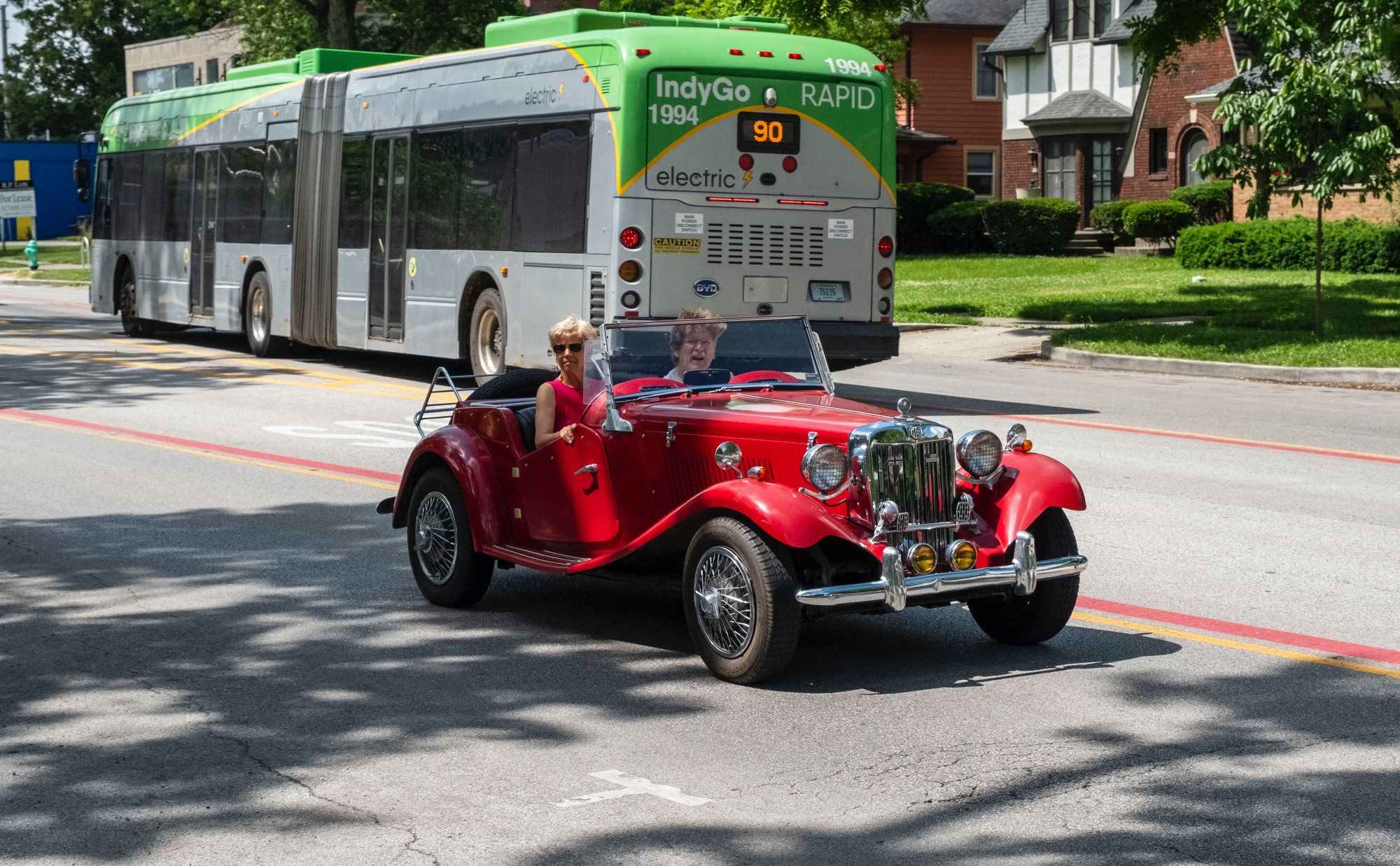 A happy-looking couple motoring up College Avenue in Indianapolis in their red MG-TD kit car on a VW Beetle chassis. They are smiling. The street is lined with trees and older homes. There is a green and silver IndyGo electric bus headed the opposite way in the background.