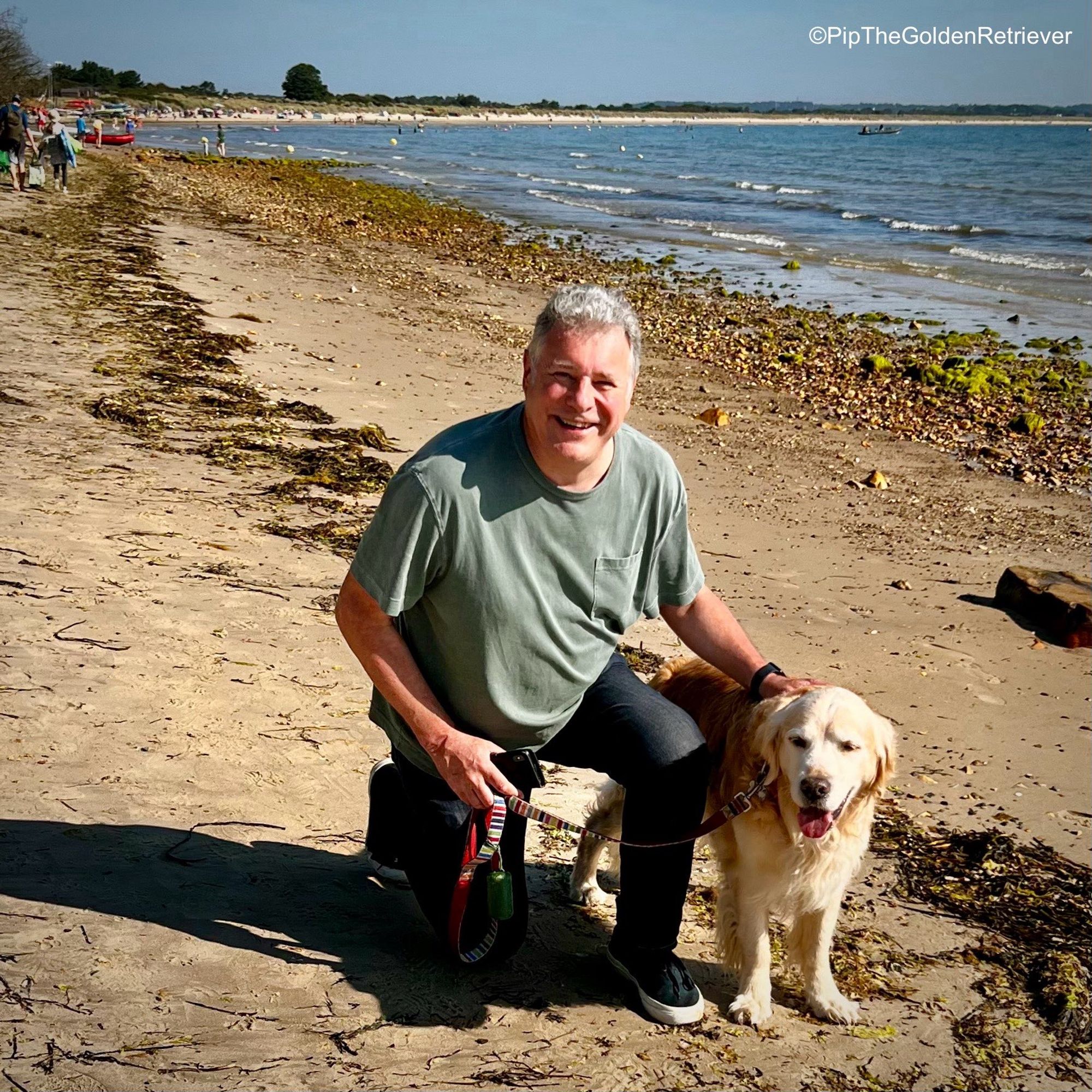 Pip the Golden Retriever is standing facing towards you on a sunny morning at Studland Bay on the Isle of Purbeck. His human father is crouched down next to him. Both are smiling at you.