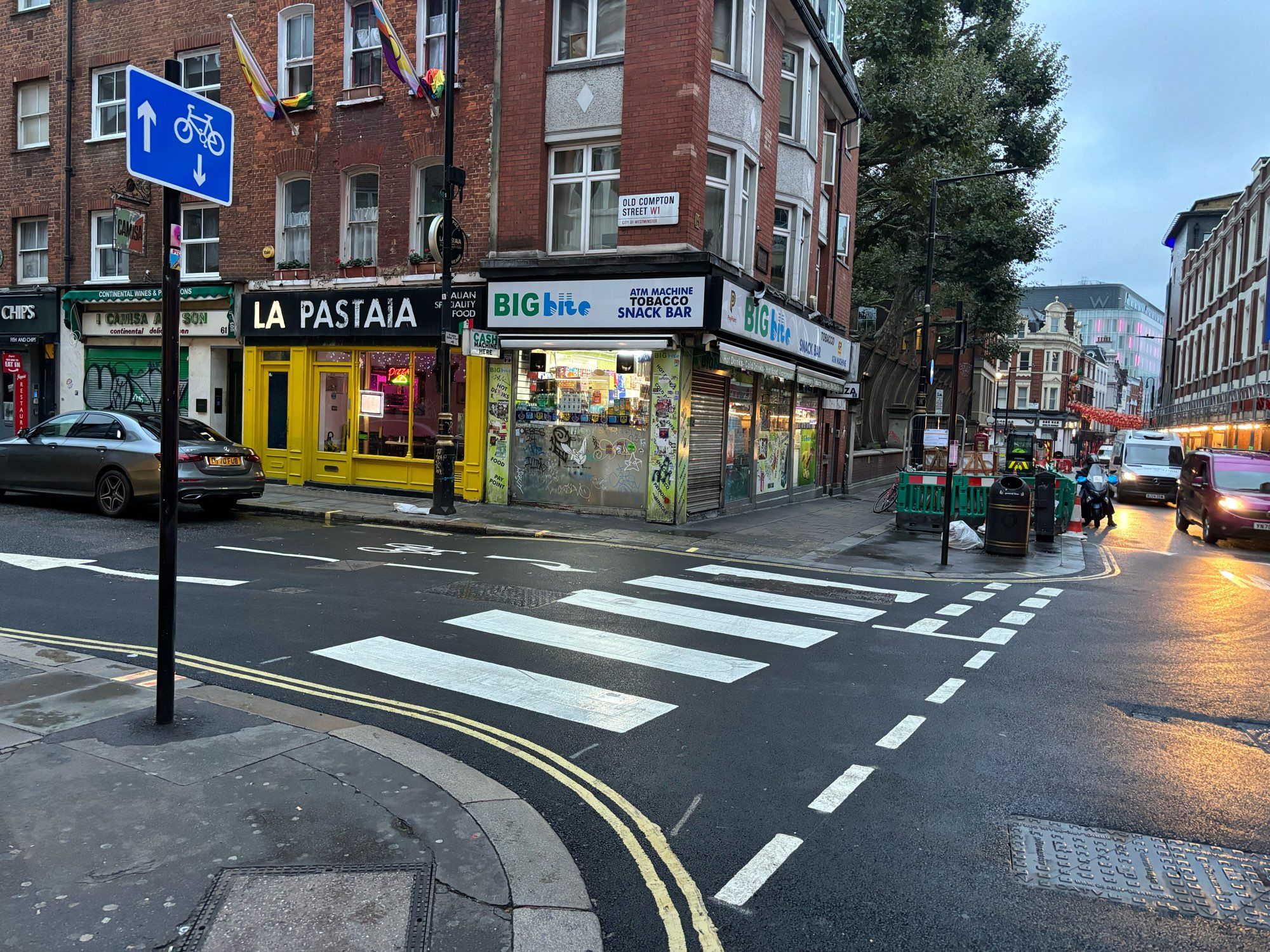 Shows a zebra crossing marked on a side road. The street is in central London. There is a shop and a cafe in the background. Vans are driving up the main road. It is damp