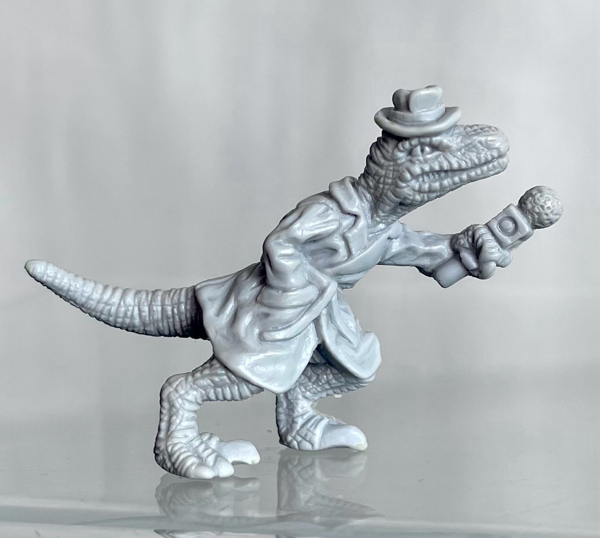 A small sculpture of a velociraptor reporter. A velociraptor reminiscent of Jurassic Park wearing a trench coat and fedora with a “press” badge, one hand in coat pocket, the other arm jutting a microphone outward.