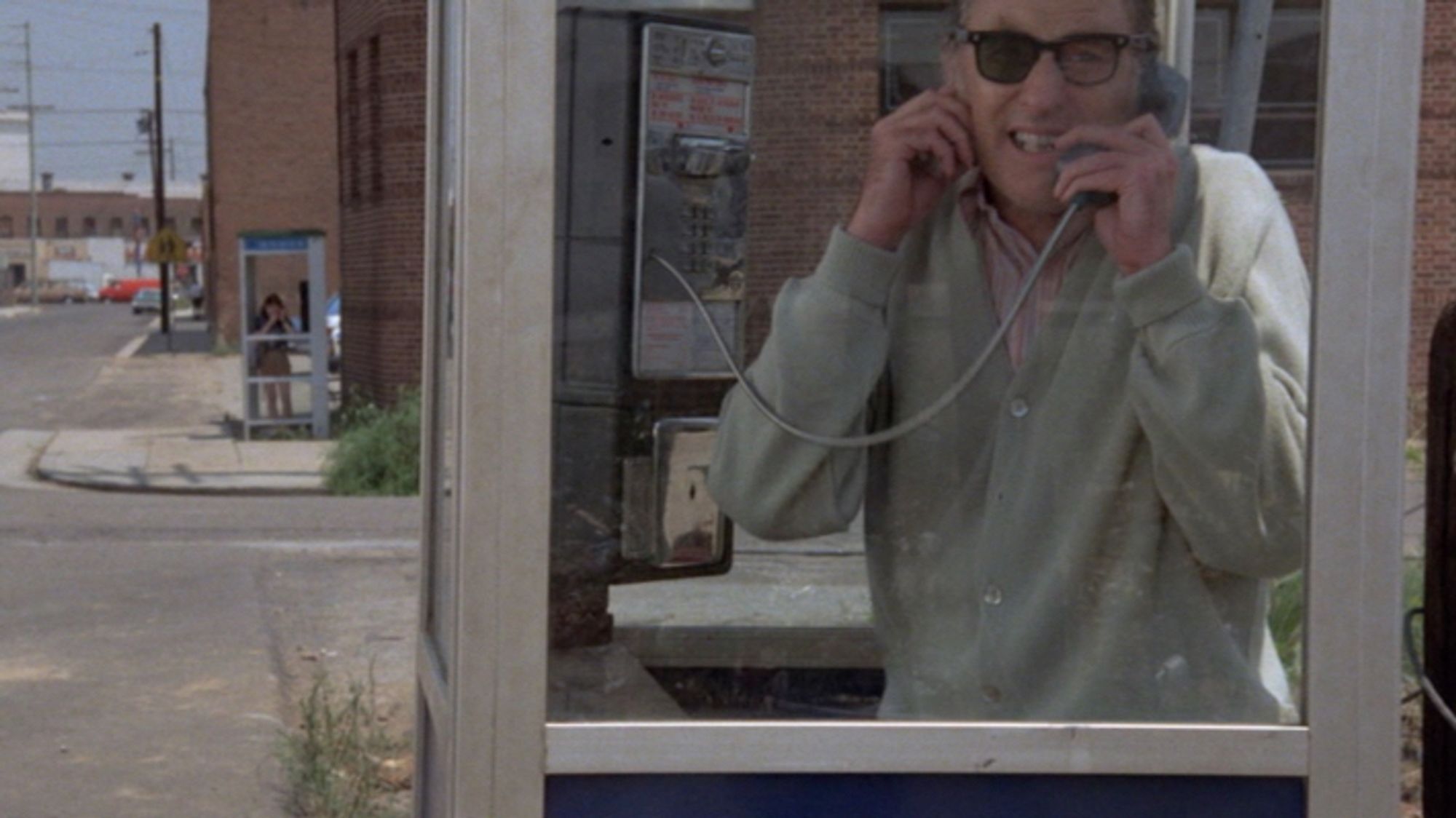 A man with half sunglasses in a phone booth talking to a woman in another phone booth across the street