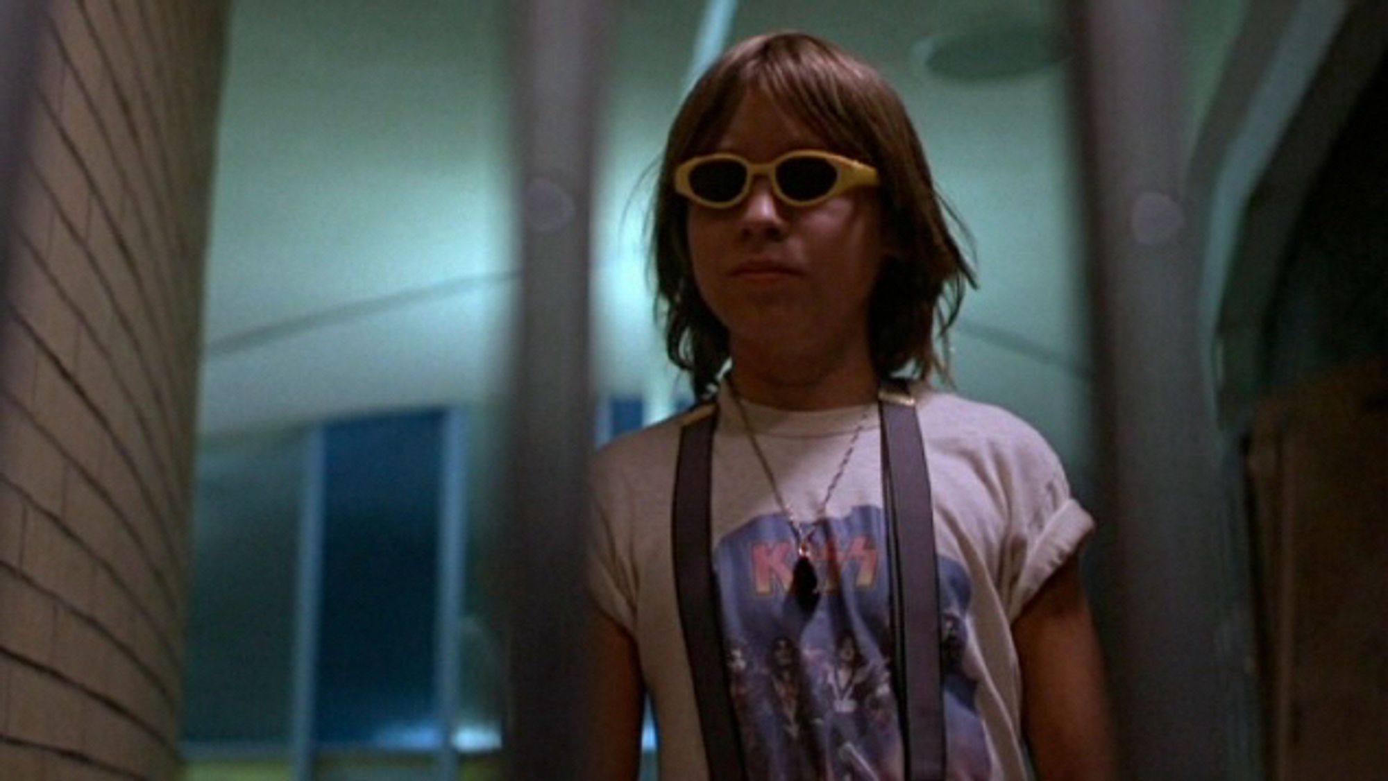 A kid with suspenders, sunglasses and a KISS shirt stands behind a gate in a school hallway