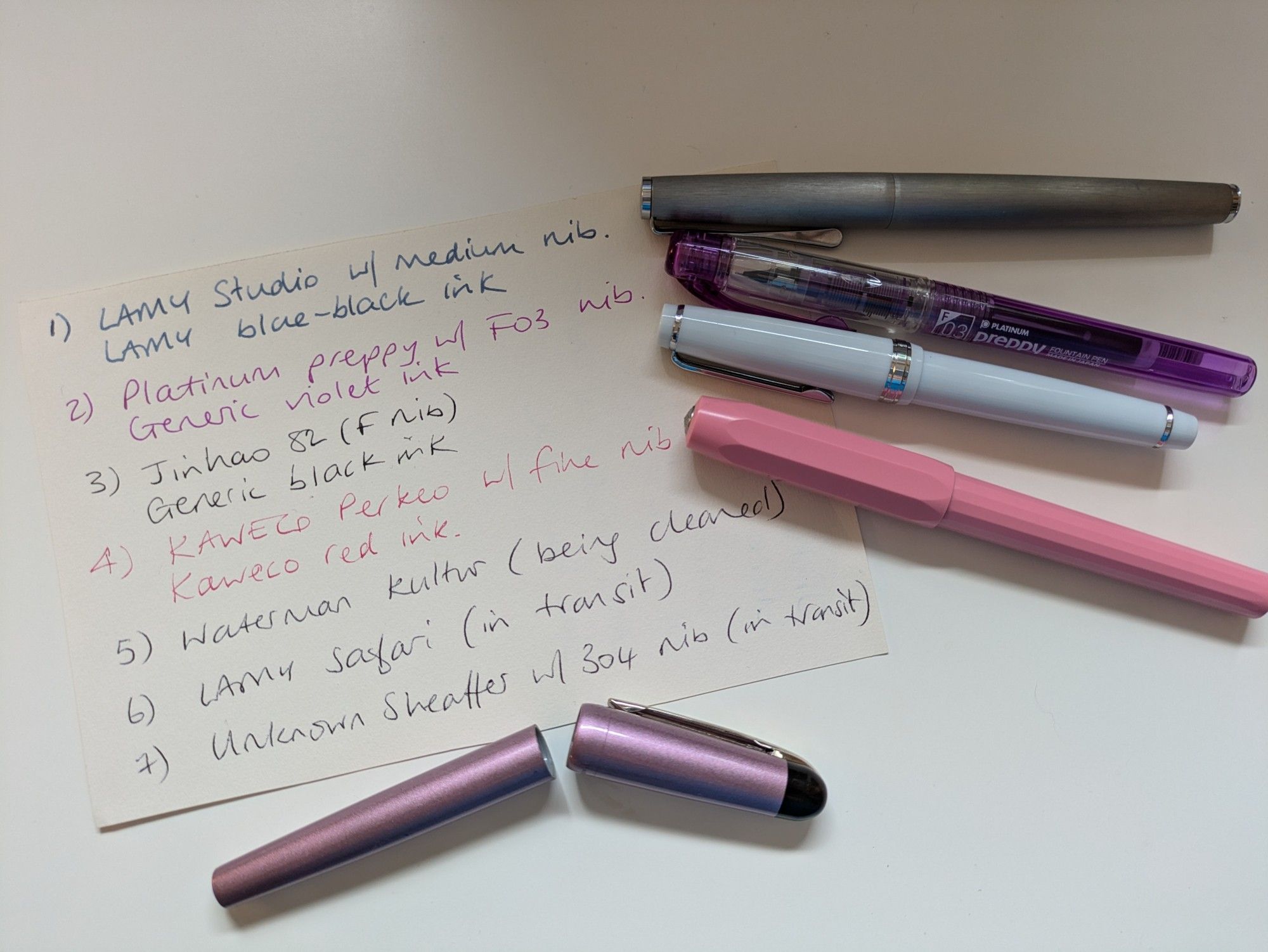 The image shows a card with a handwritten list of pen names and inks, to the right are four fountain pens (LAMY studio in brushed steel, Platinum Preppy in purple, Jinhao 82 in pale blue, Kaweco Perkeo in peony pink). To the bottom of the image is the outer casing of a Waterman Kultur pen which is currently having it's nob cleaned.