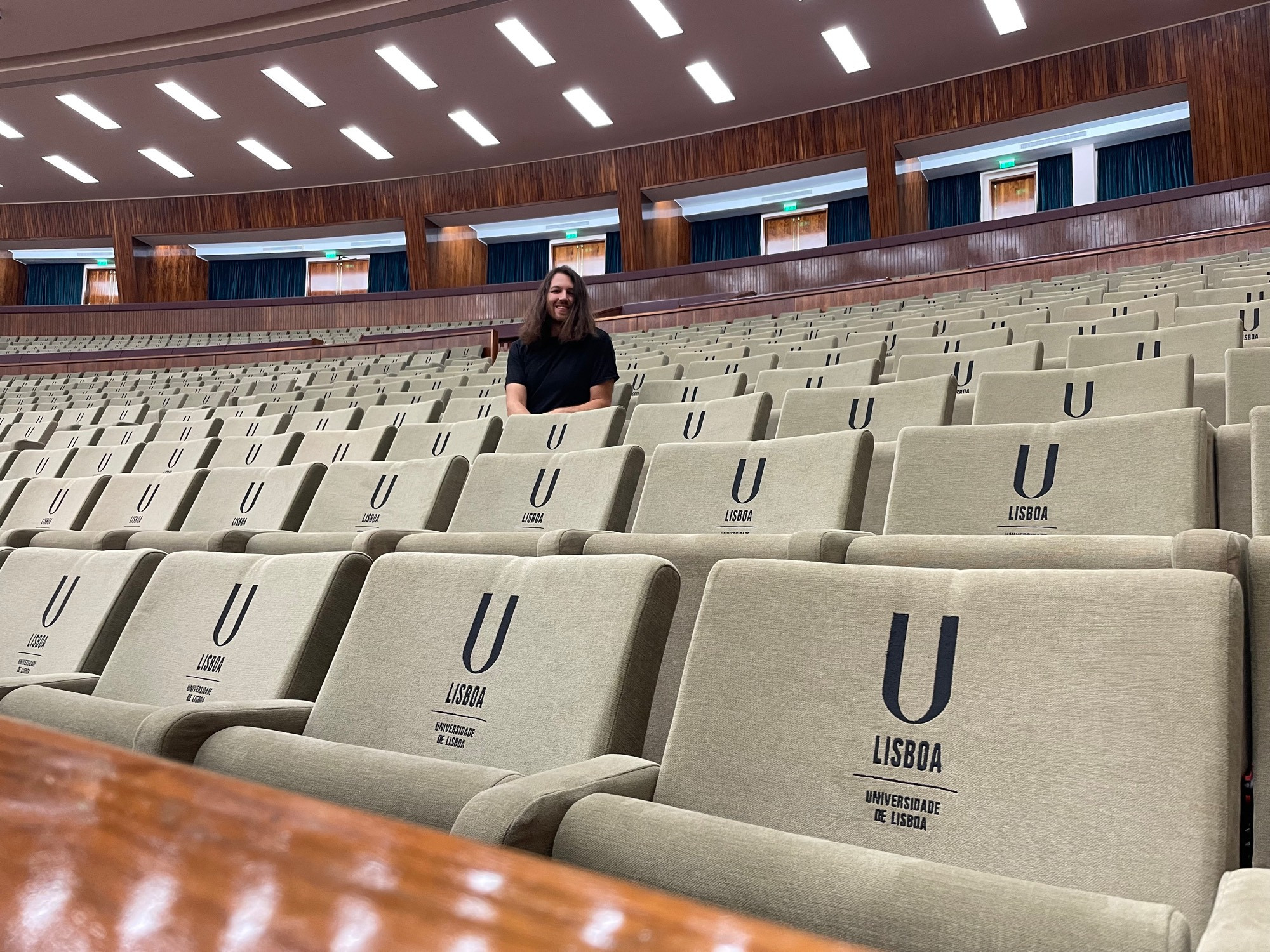 Person in the middle-distance sitting in a mid-century auditorium, occupying one of uncountable mint-ish green chairs, all of which are embroidered with “U Lisboa”