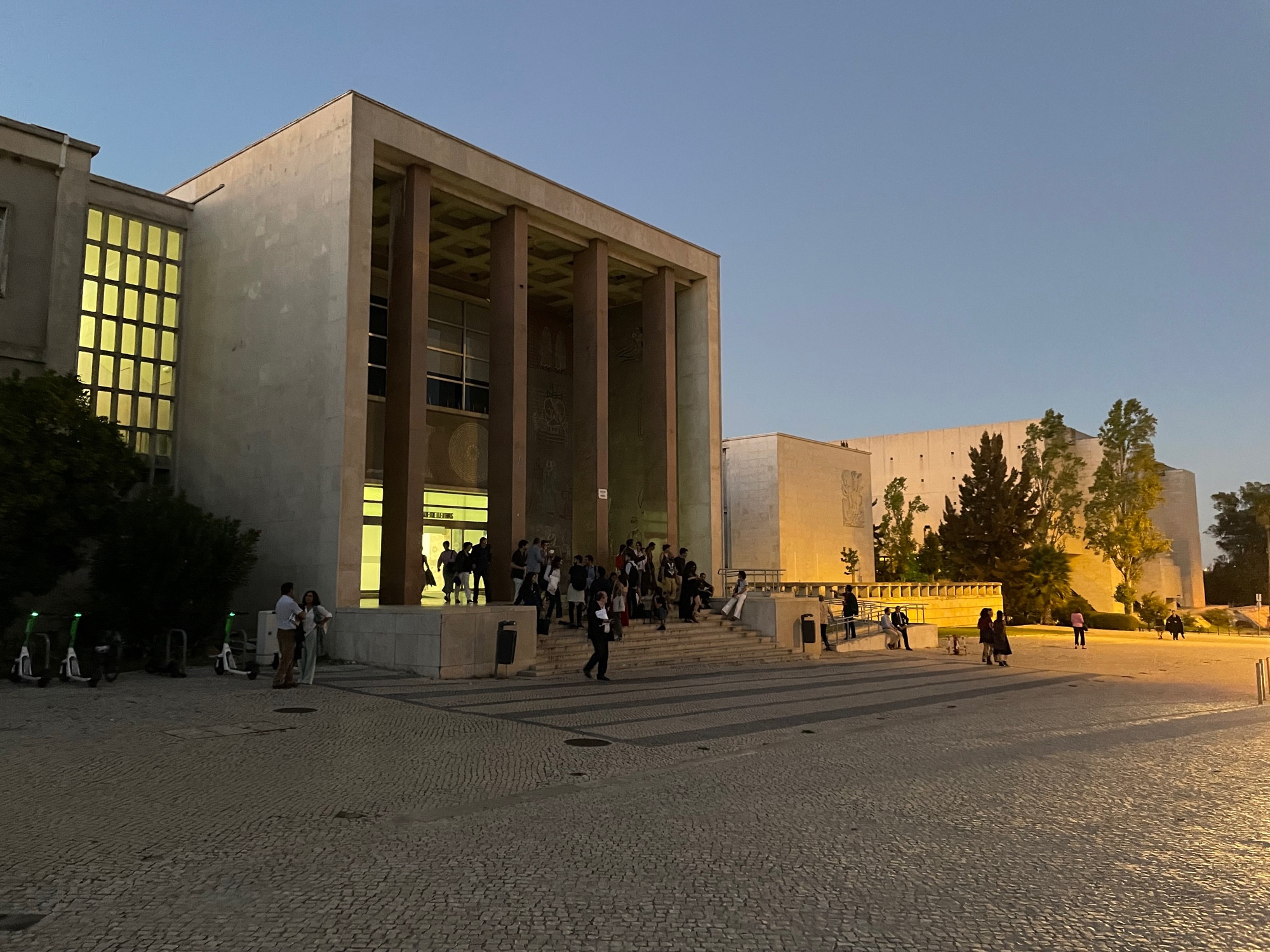 A late-50s university building entrance with huge, rectangular columns in the early evening, groups of people sit and stand in front of it.