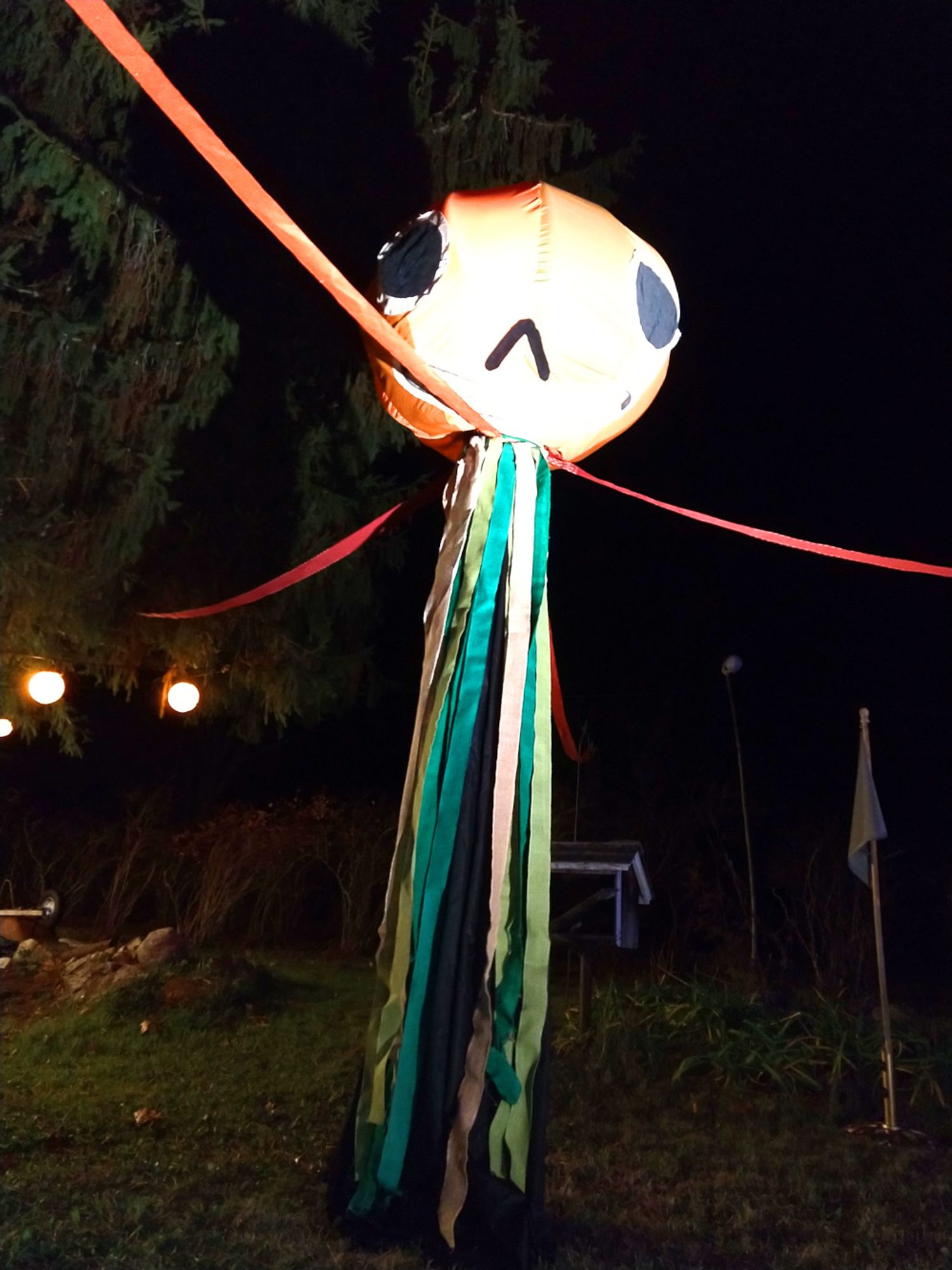 An image of a life size Enoch Halloween prop. Standing at over 13 feet tall, Enoch is like a May Pole, but with a giant cloth jackolantern head on the top. Large black eyes, a chevron-shaped black nose hole, and a toothy grin adorn the large, jackolantern head. His body is thin and made up of ribbon streamers, four of which stretch out like great, long arms, while the rest dangle like spindly legs.