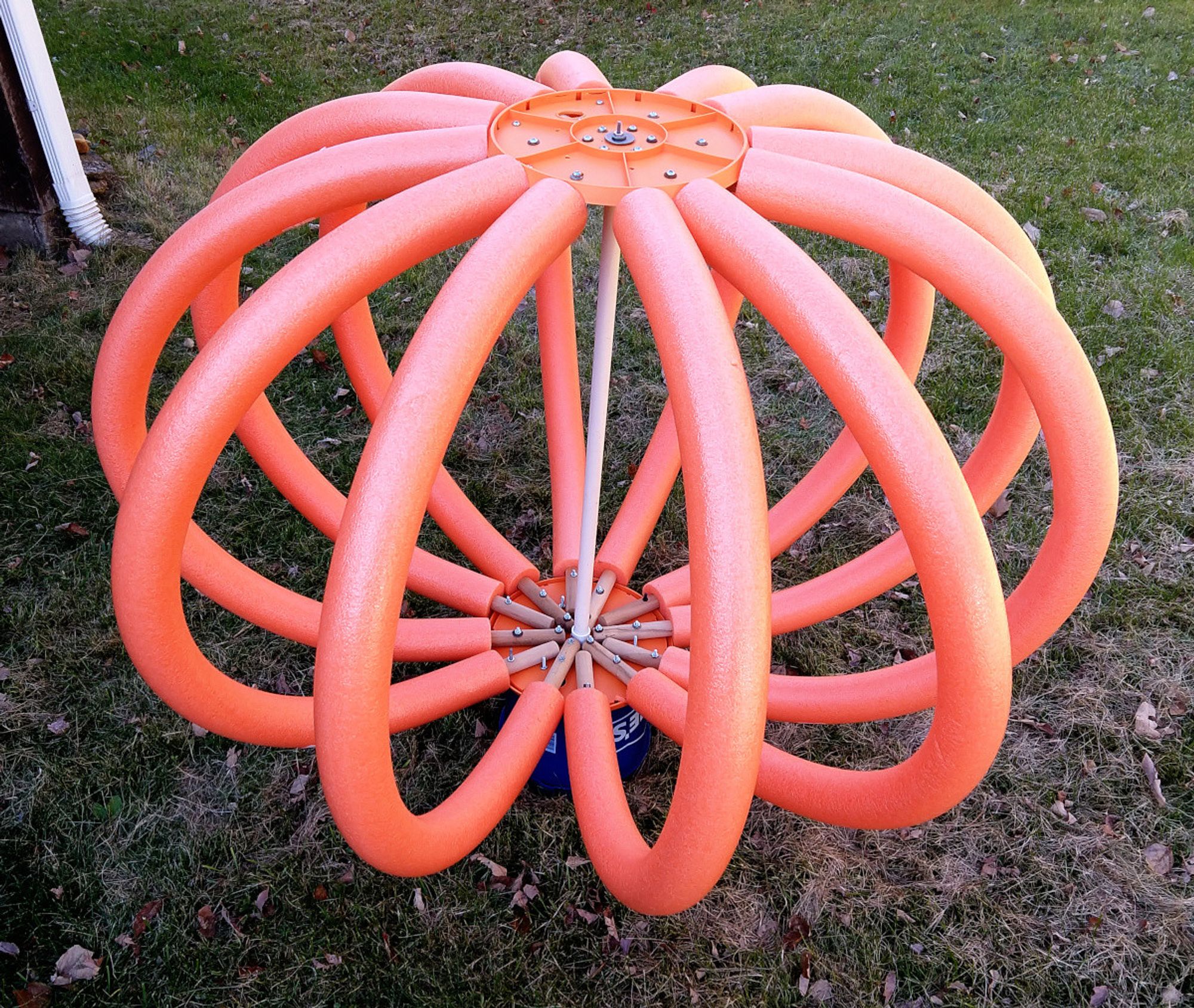 An image of the interior of the head of my life size Enoch prop, which consists of twelve orange pool noodles arching between top & bottom rows of radial wooden dowels attached to plastic circles. The internal structure is supported by a central piece of PVC pipe. The bottom is attached to an upside-down bucket, which attaches to his base: a folded up clothes line.