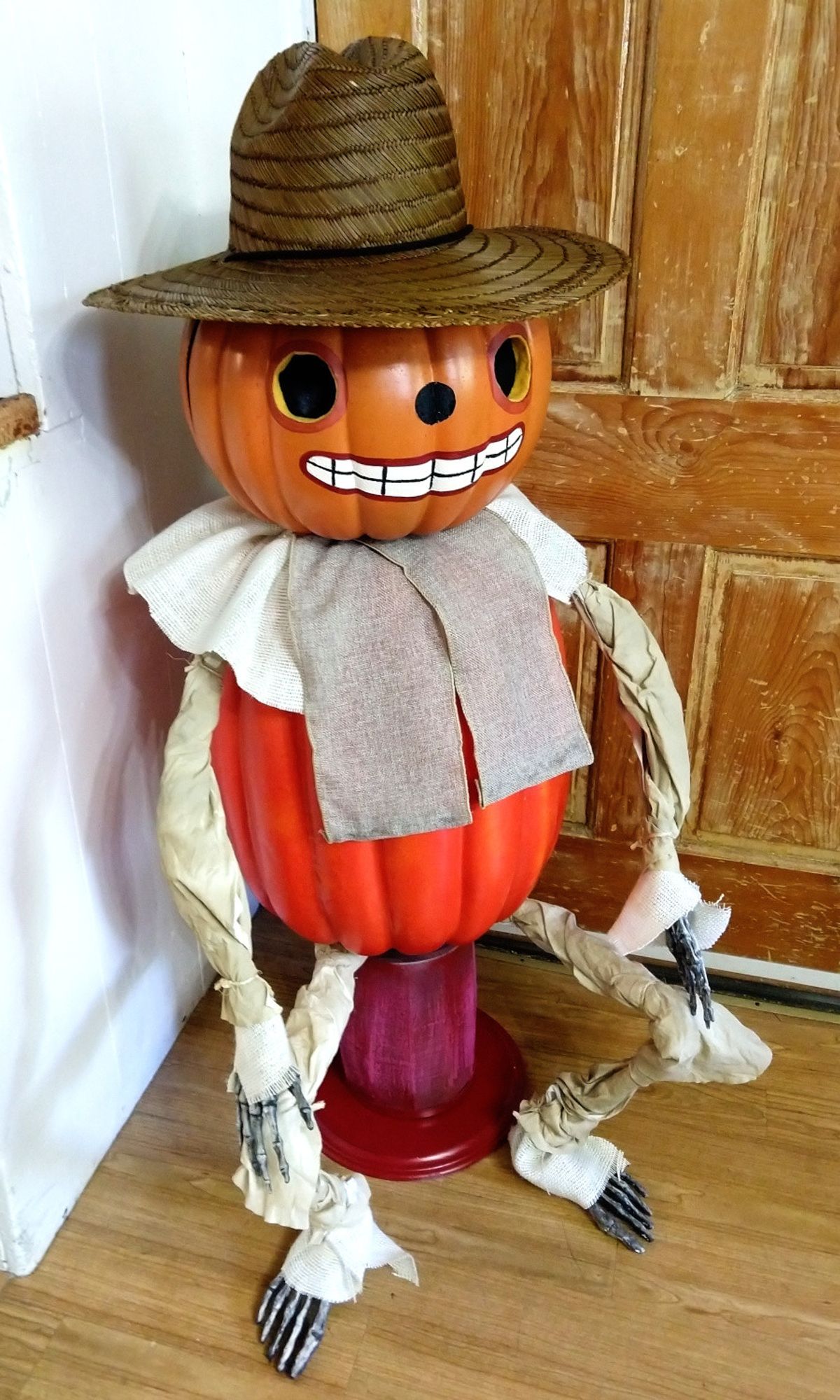 An image of a life size Potsfieldian from Over The Garden Wall. A skeleton wearing pumpkins on their torso & head, with the head carved and painted with a smile, corn husks covering the arms and legs, wearing a woven grass sun hat and sitting on a small red stool.