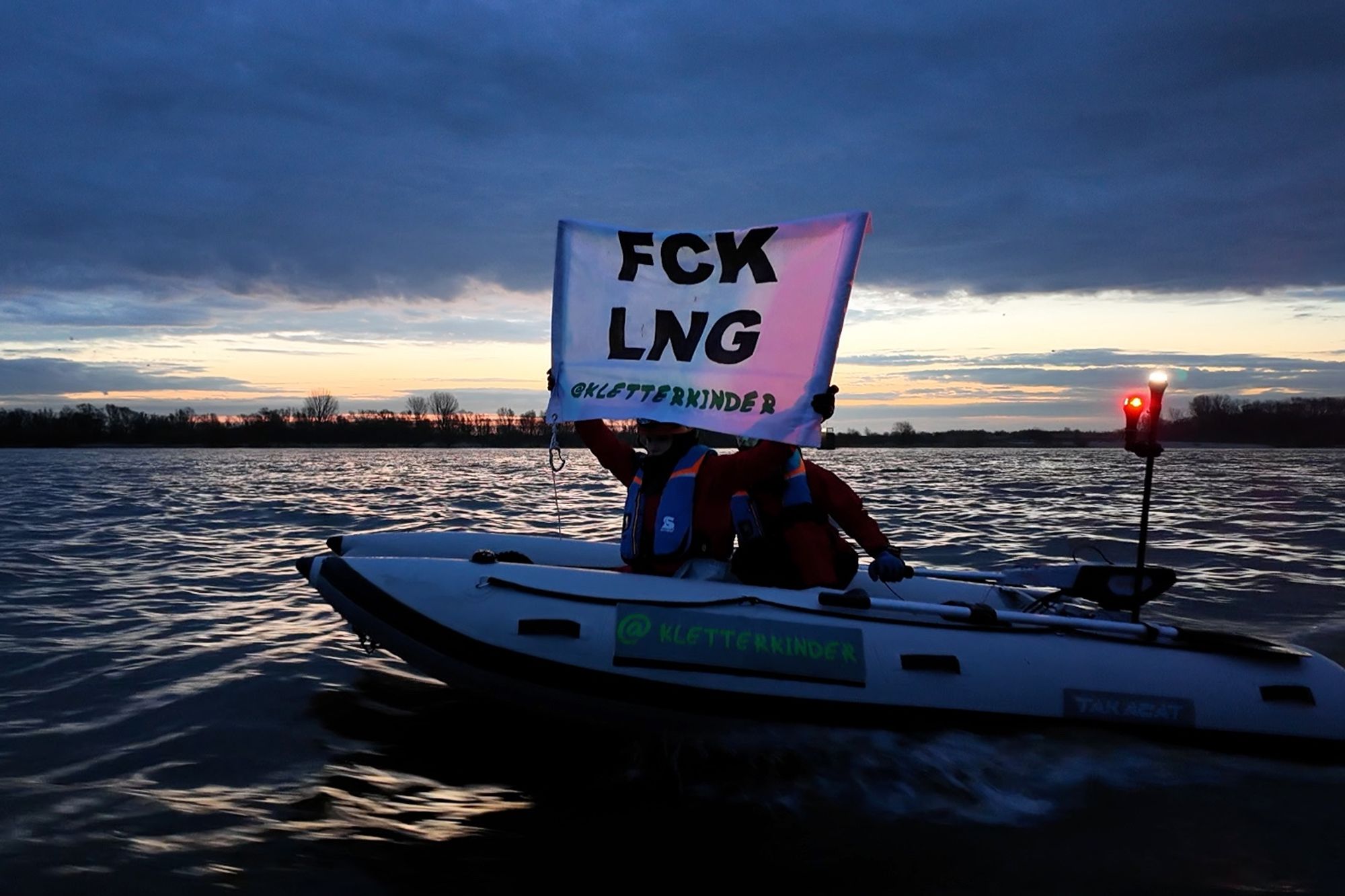 Photography of the two Kletterkinder siblings in their E-powered inflatable catamaran on river Elbe near Stade, holding a banner with inscript “FCK LNG” (you can guess what that means, right? Hint: LNG stands for Liquefied Natural Gas. (© Kletterkinder)