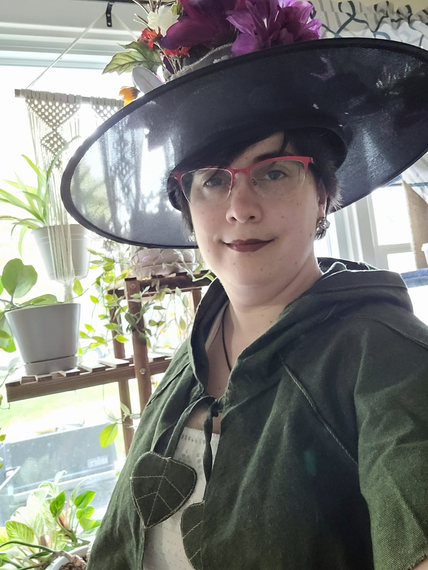 A witchy selfie of Redria. Houseplants line a window shelf behind them. She has a black witch hat with a floral arrangement, a white dress, and a green leafy capelet