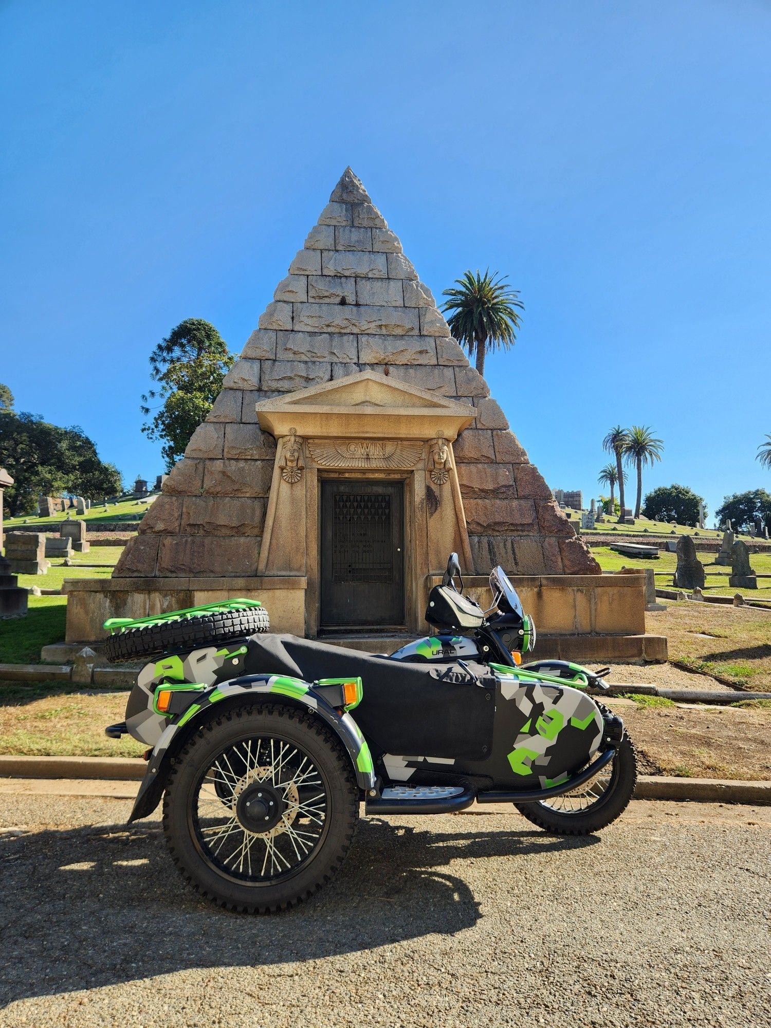 A Ural GearUp in green/black digicam parked in front of a pyramid mausoleum at a cemetery in Oakland, CA.