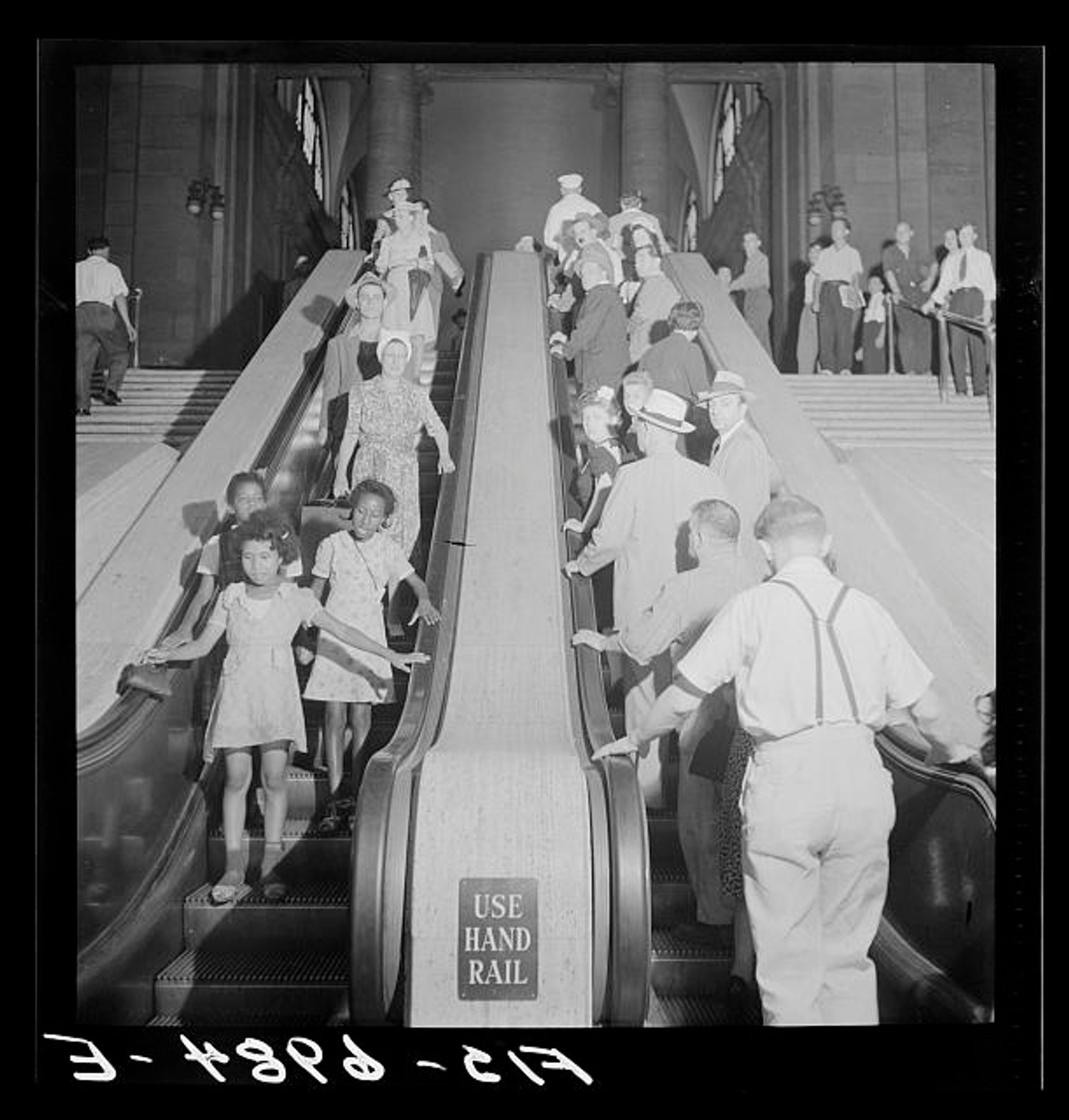 African American children and white adults ride the escalators in two different directions in a black and white photo.