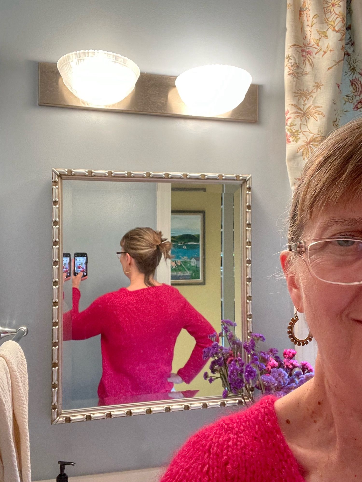 Knitter wearing raspberry-colored knitted sweater, taking photograph of the fit in a bathroom mirror