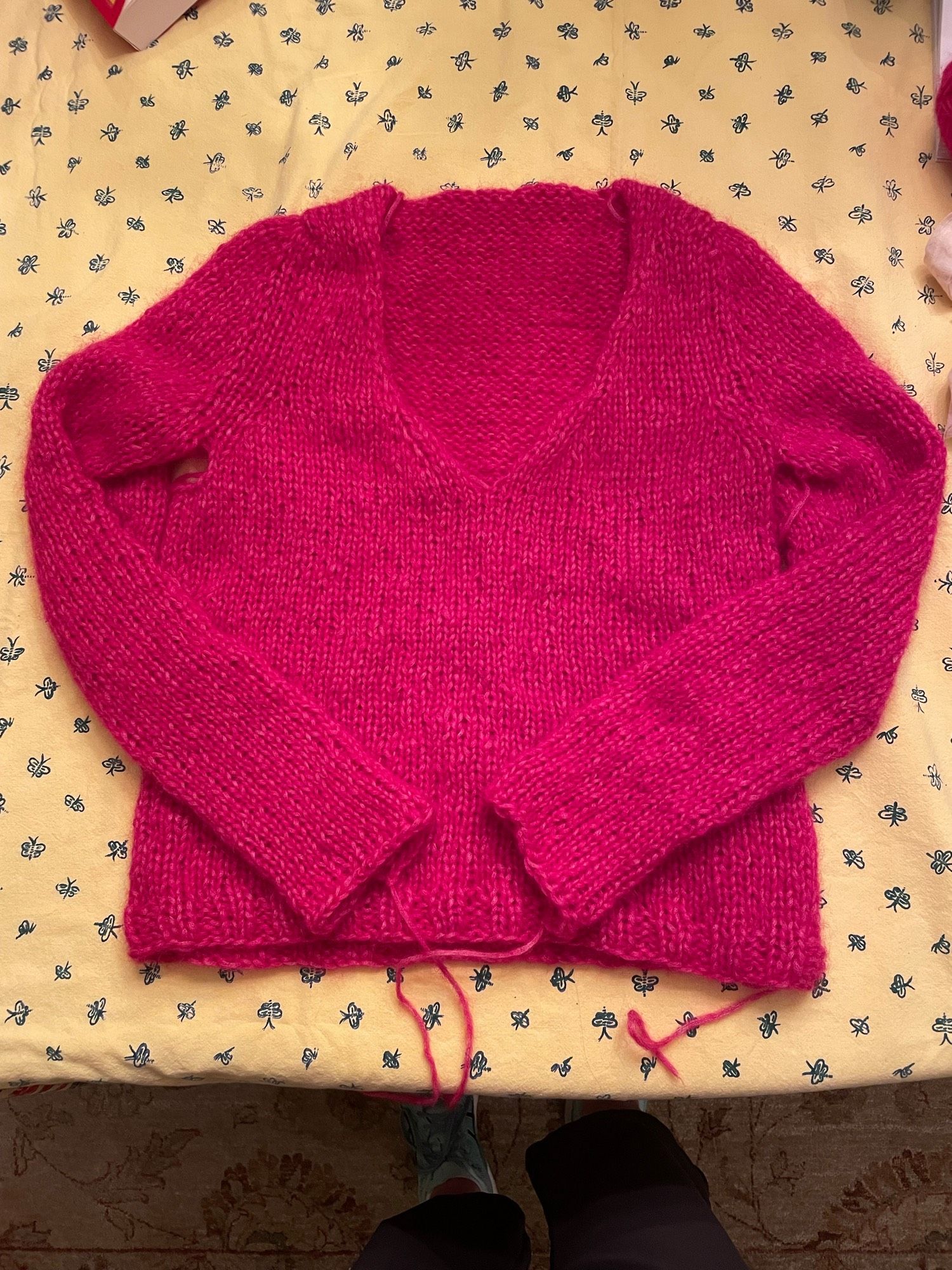 Bright raspberry-colored hand-knitted vee-neck sweater with long sleeves and a few loose ends, laid out on a yellow and blue tablecloth