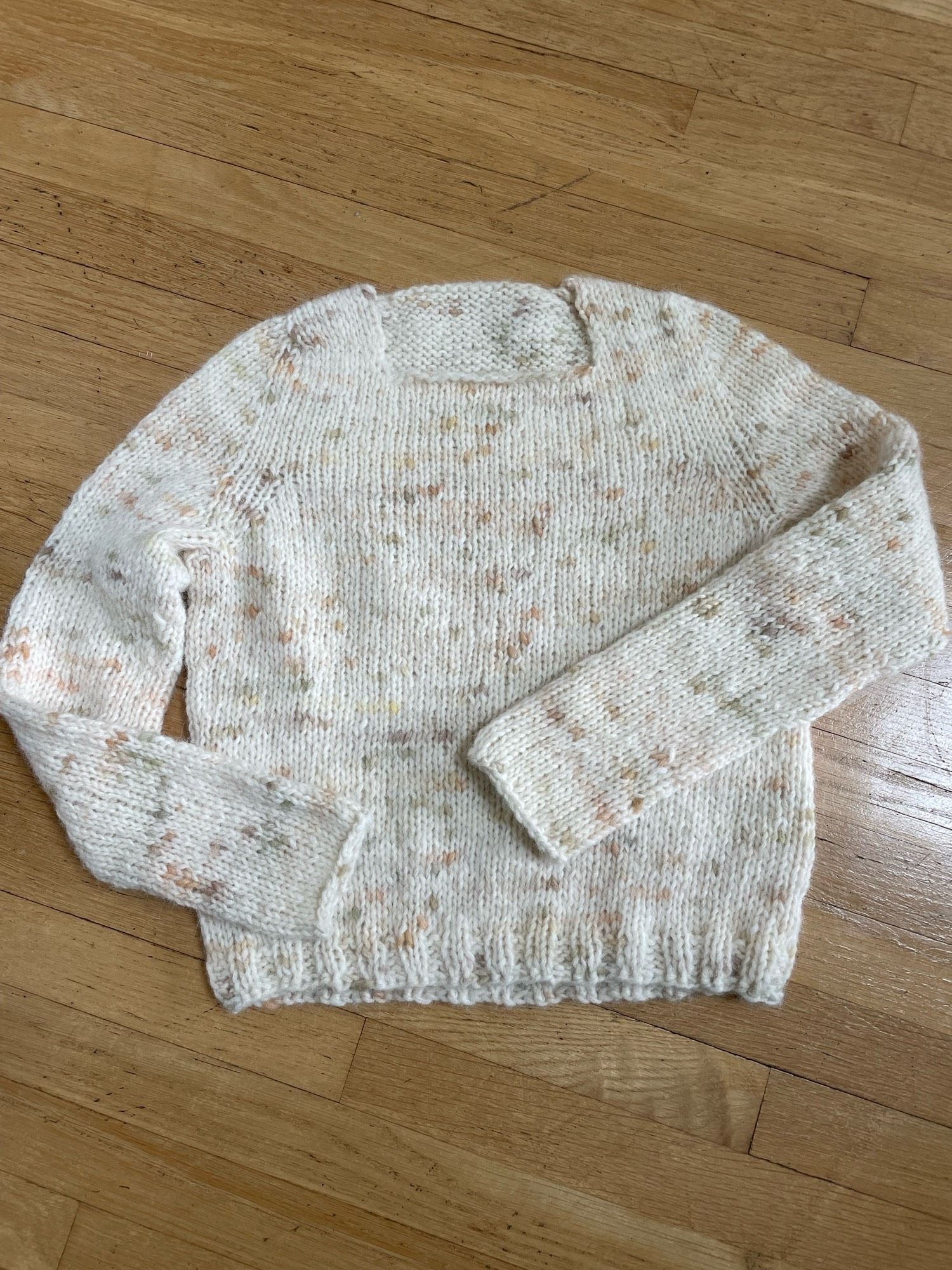 Hand-knitted long sleeve sweater in cream wool with beige, gold, and green flecks, laid out on wooden floor