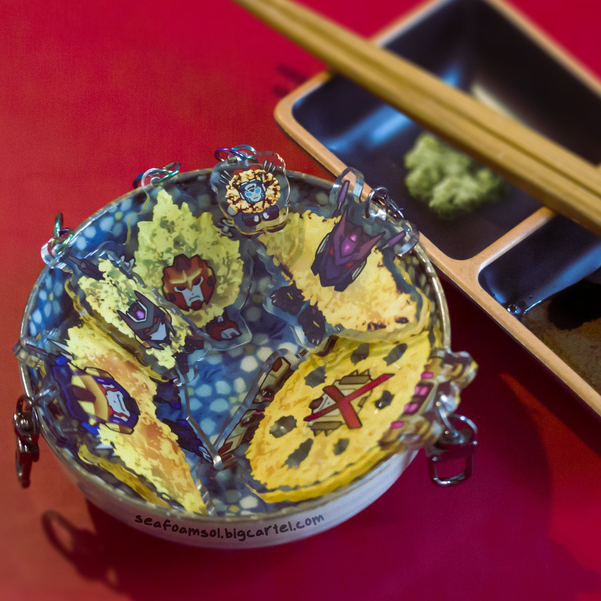 A photo of several tempura-themed keychains placed in a shallow, decorative dish. In the background, chopsticks lie across another dish in the background, which holds soy sauce and wasabi.