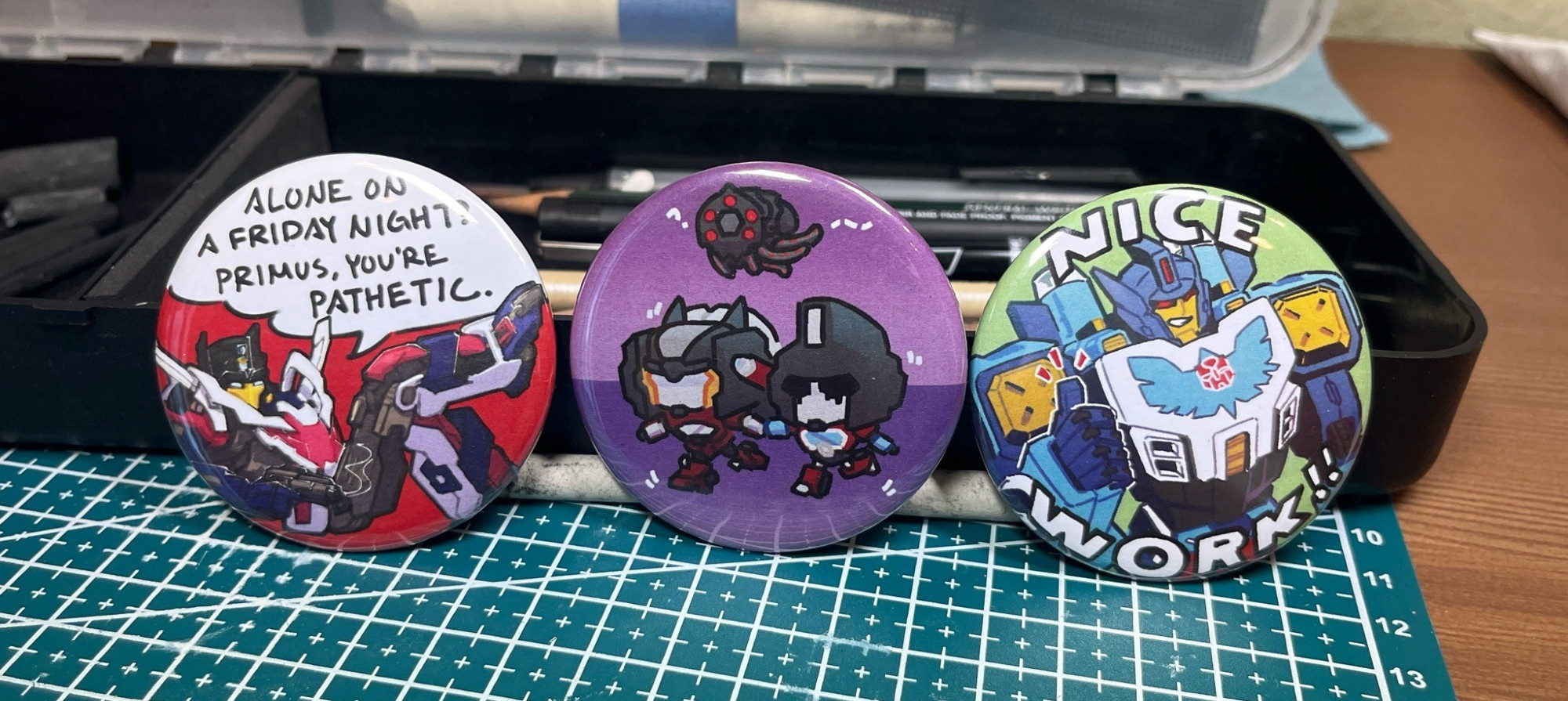 A photo of three 2.25inch buttons. They depict, from left to right, an IDW Getaway parody of the sonic "alone on a Friday night?" meme, Cyberverse chibi Dead End and Perceptor holding hands as they run from a quintesson prosecutor, and IDW Thunderclash holding a thumbs up saying "nice work!!"