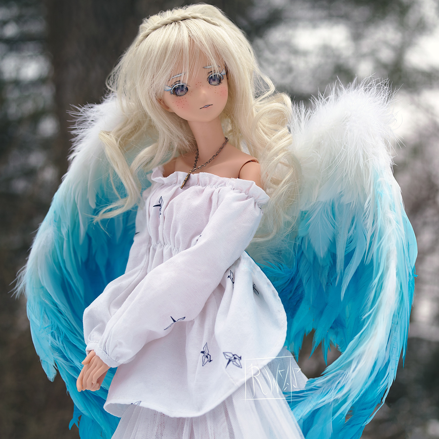 A ball-jointed doll with blonde hair and blue, feathery wings stands in the middle of the frame. She is wearing a white dress with a pattern of origami cranes on it. She is looking to the right.