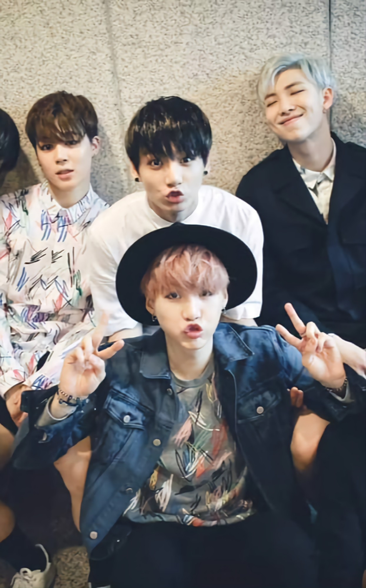 Yoonkook, Jimin, and Namjoon sitting down, posing for the camera. Jungkook is in a white shirt with black plugs on his ears. Suga is in a denim jacket over a fancy shirt and he's got in a black hipster hat.