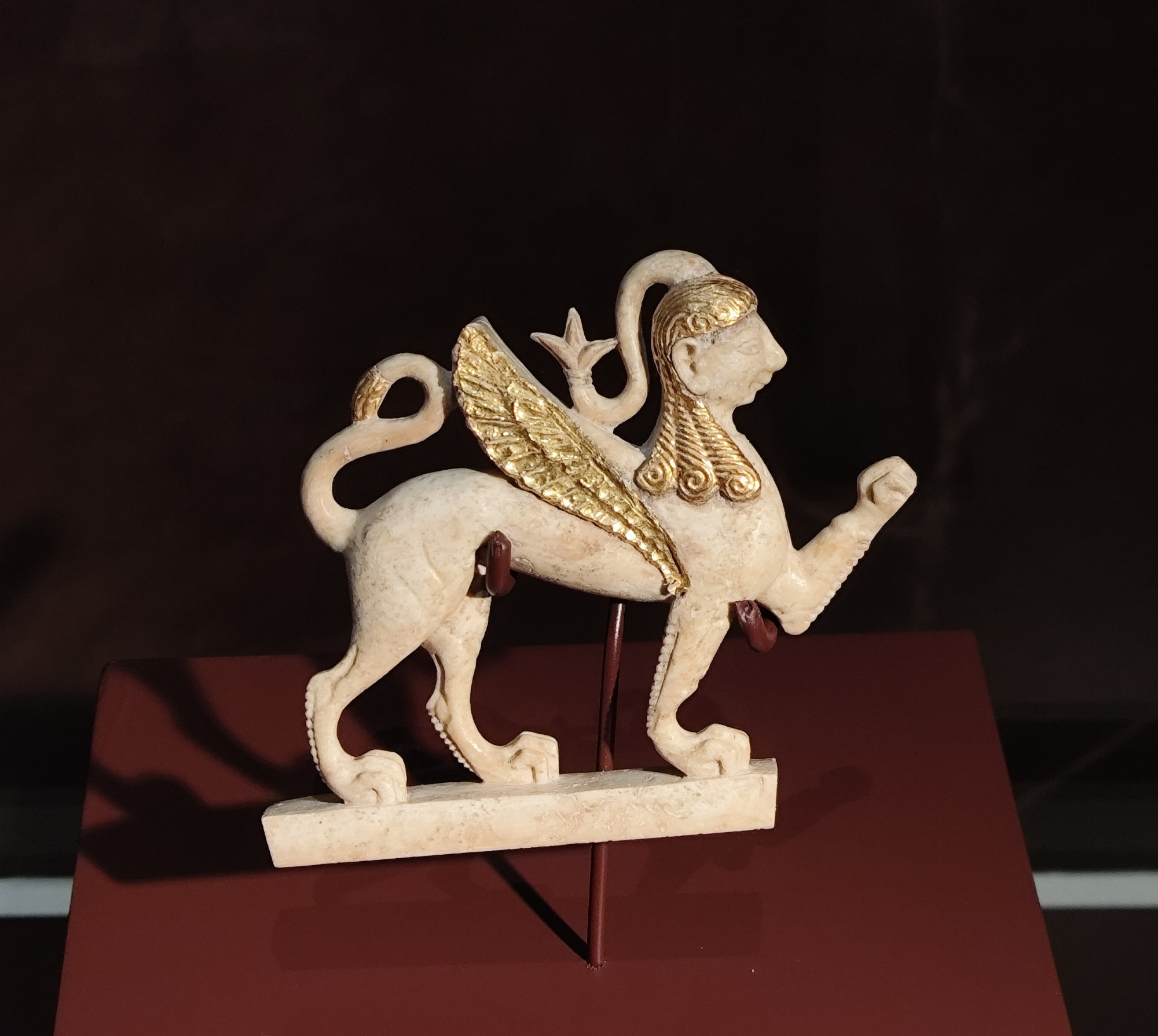 A sphinx on four feet, one of the forefeet is extended forward. Ivory, wing and hair is gold coated