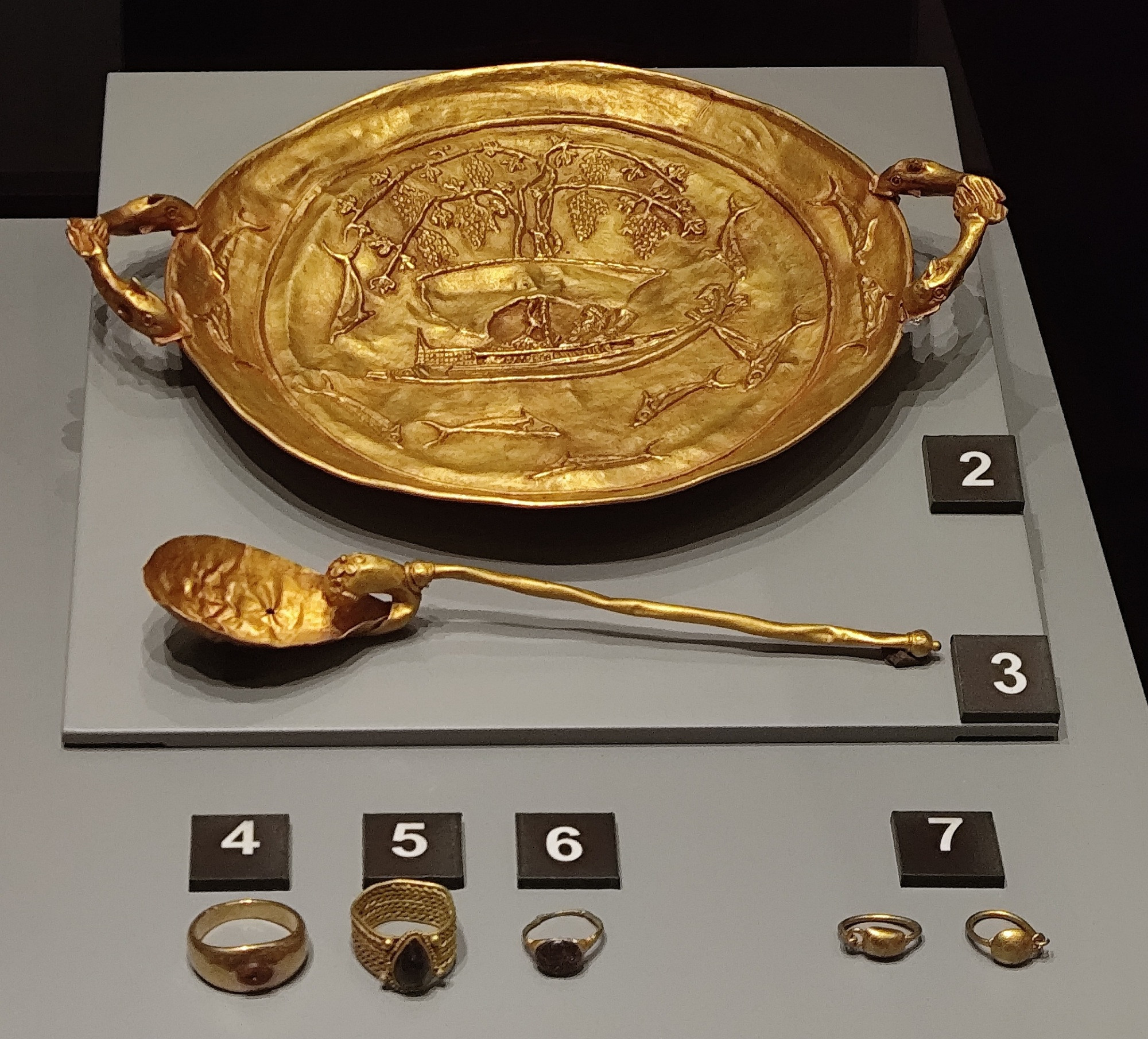 A round golden tray, on it is a boat going under a vine with big grape bunches. Under it, dolphins are swimming. Each of the 2  handles were made of two dolphins attached mouth to mouth. Next to the tray was placed a golden spoon with  lion protome on the attachment of the handle. Next to them, 3 golden gemstone rings and a pair of golden earrings.
