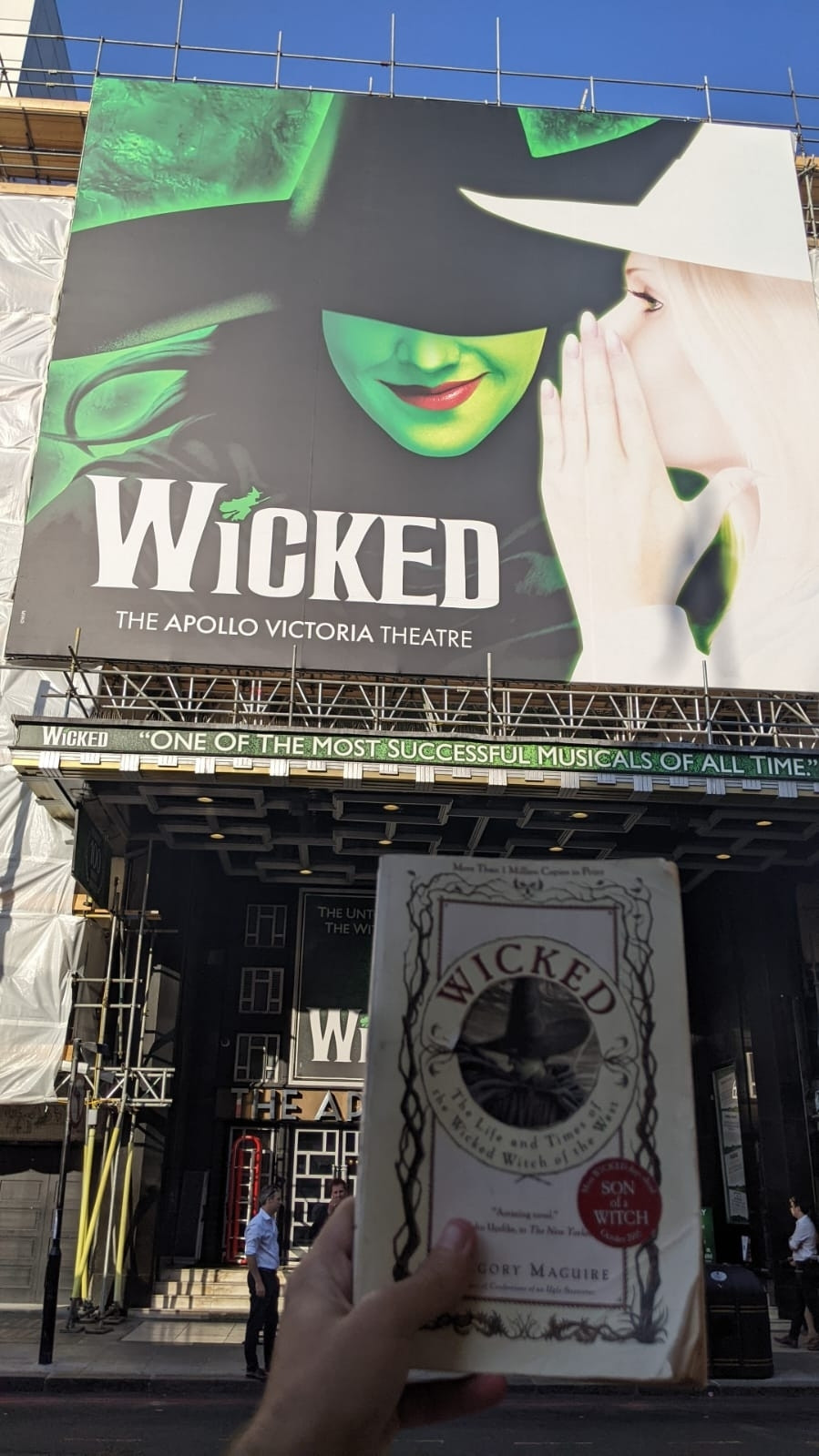 The big billboard of Wicked musical showing a green person with black hair, wearing a witch's hat, smiling (we just see her smile) while a person dressed in white clothes and hair whisper a secret.
This billboard is in the front of the theater, a hand is holding a book from up close which is also called wicked