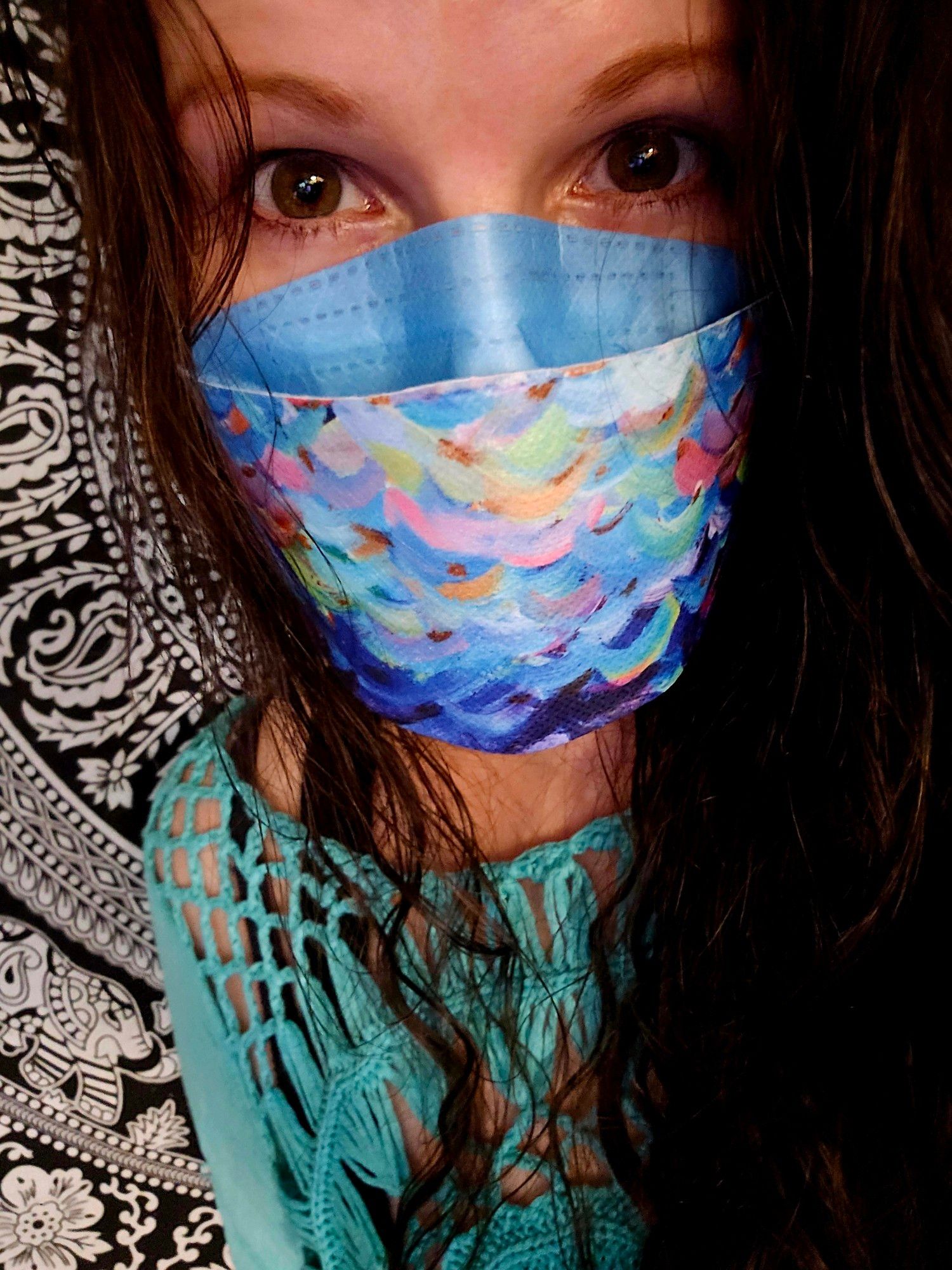 Female wearing @masklab blue painted mask over aura n95 with turquoise knit top and black and white mandala tapestry in background.