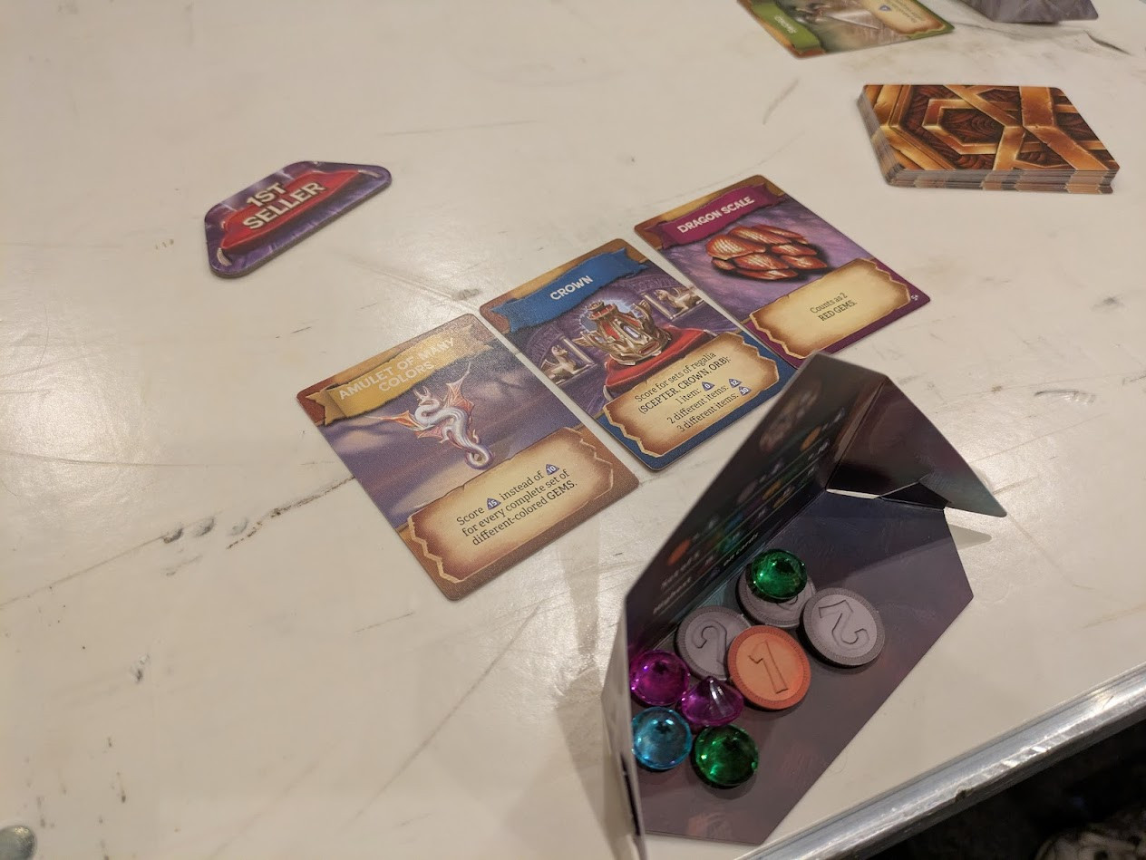 A screen hiding gems and coins. Cards laid out in front of the screen. A token that says "1st Seller"