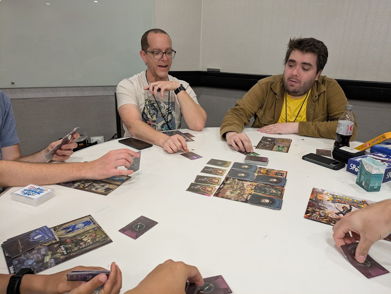 People sitting at a table, each with a hidden card about to be revealed. A board in the center showing zombies.
