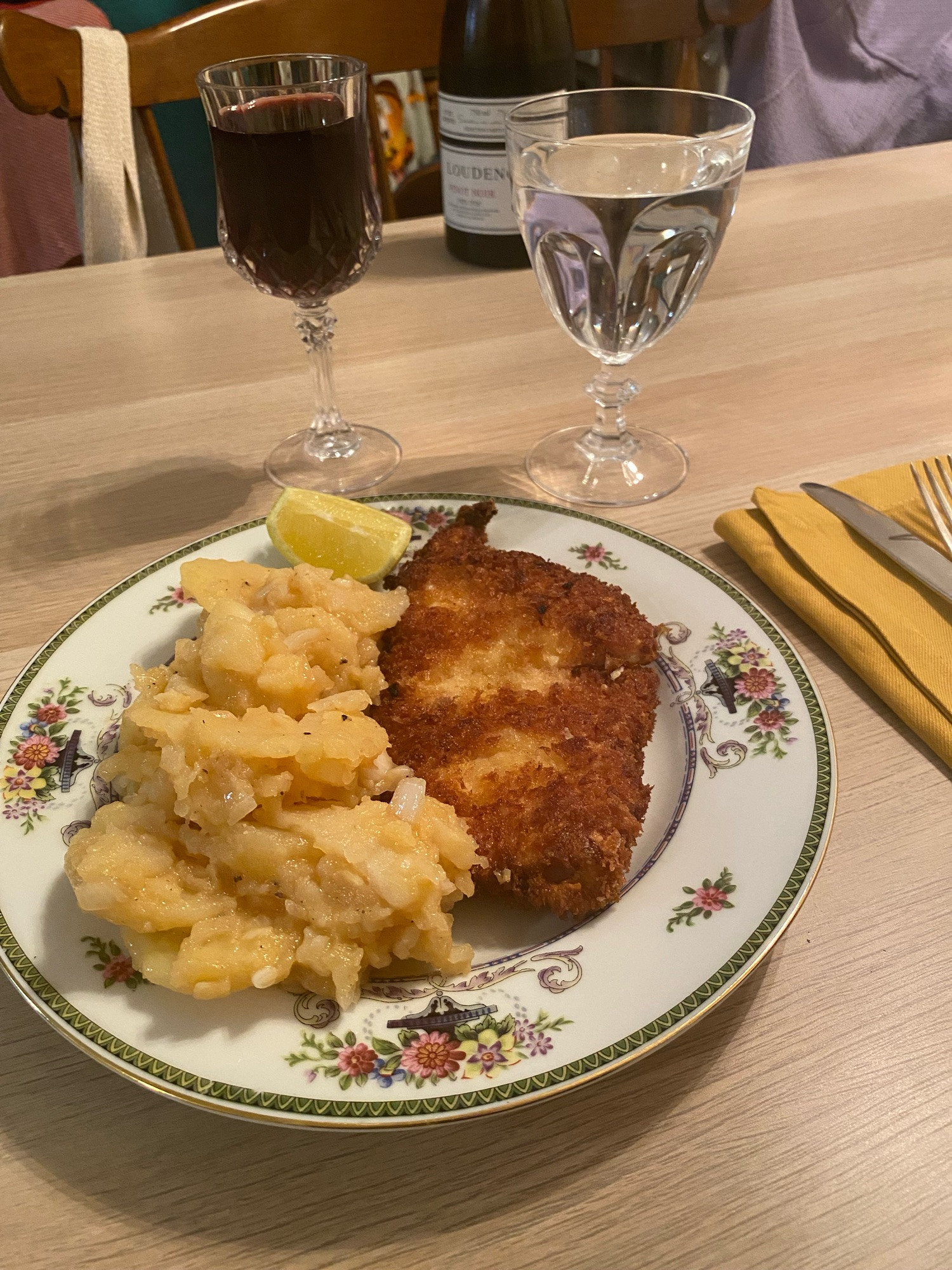 A plate with a chicken schnitzel and German potato salad, glasses of water and Pinot noir behind it