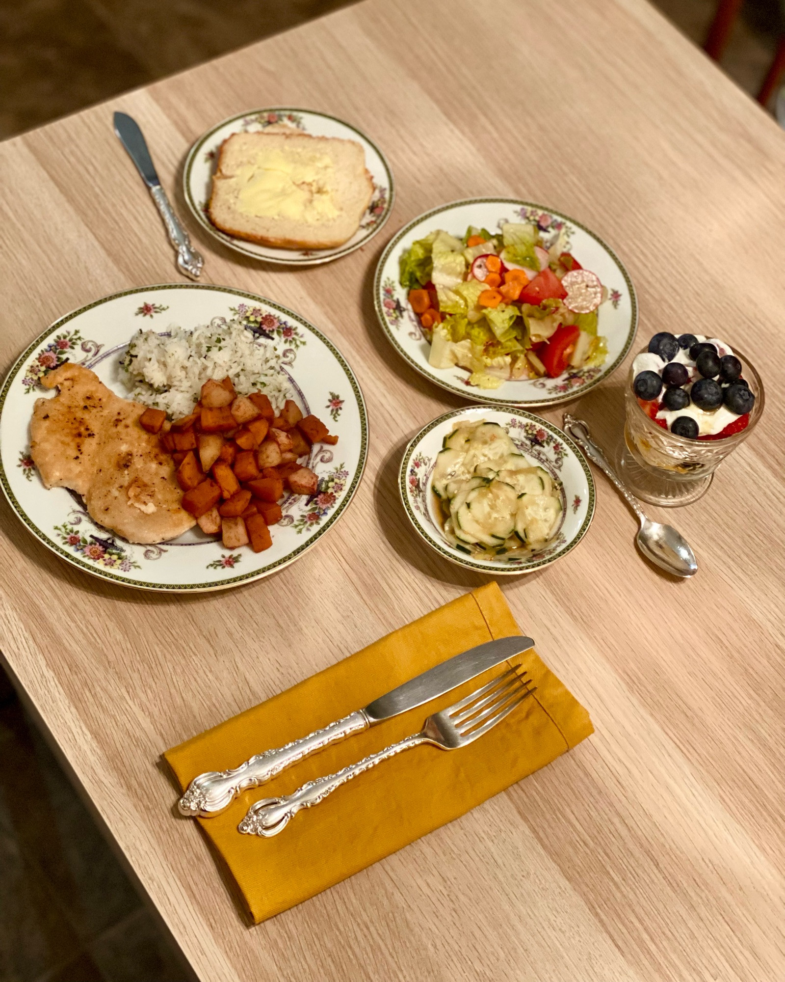 Dinner, consisting of a plate with a thin cut chicken breast, rice seasoned with furikake, sweet sweet potatoes, a cucumber salad, a green mixed salad, two buttered slices of bread, and a fruit parfait with sour cream and honey.