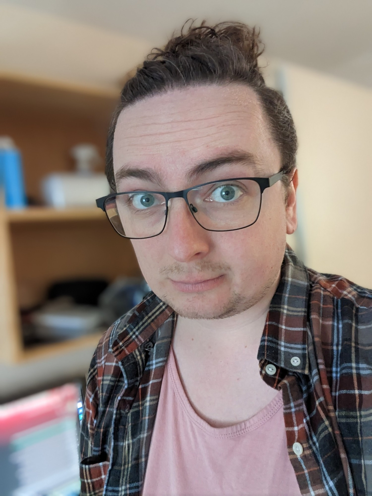 Awkward selfie of a young male-presenting human with a autumnal red/grey plaid shirt over a pink undershirt. He has Brown hair that is greying and blue eyes. He wears black square rimmed glasses. 