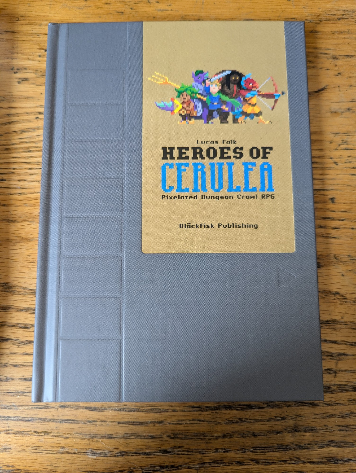 A gray book with a brown-gold label reading "Heroes of Cerulea". Above the name there is a grouping of pixel art characters. The cover of the book is stylized to look like an NES cartridge.