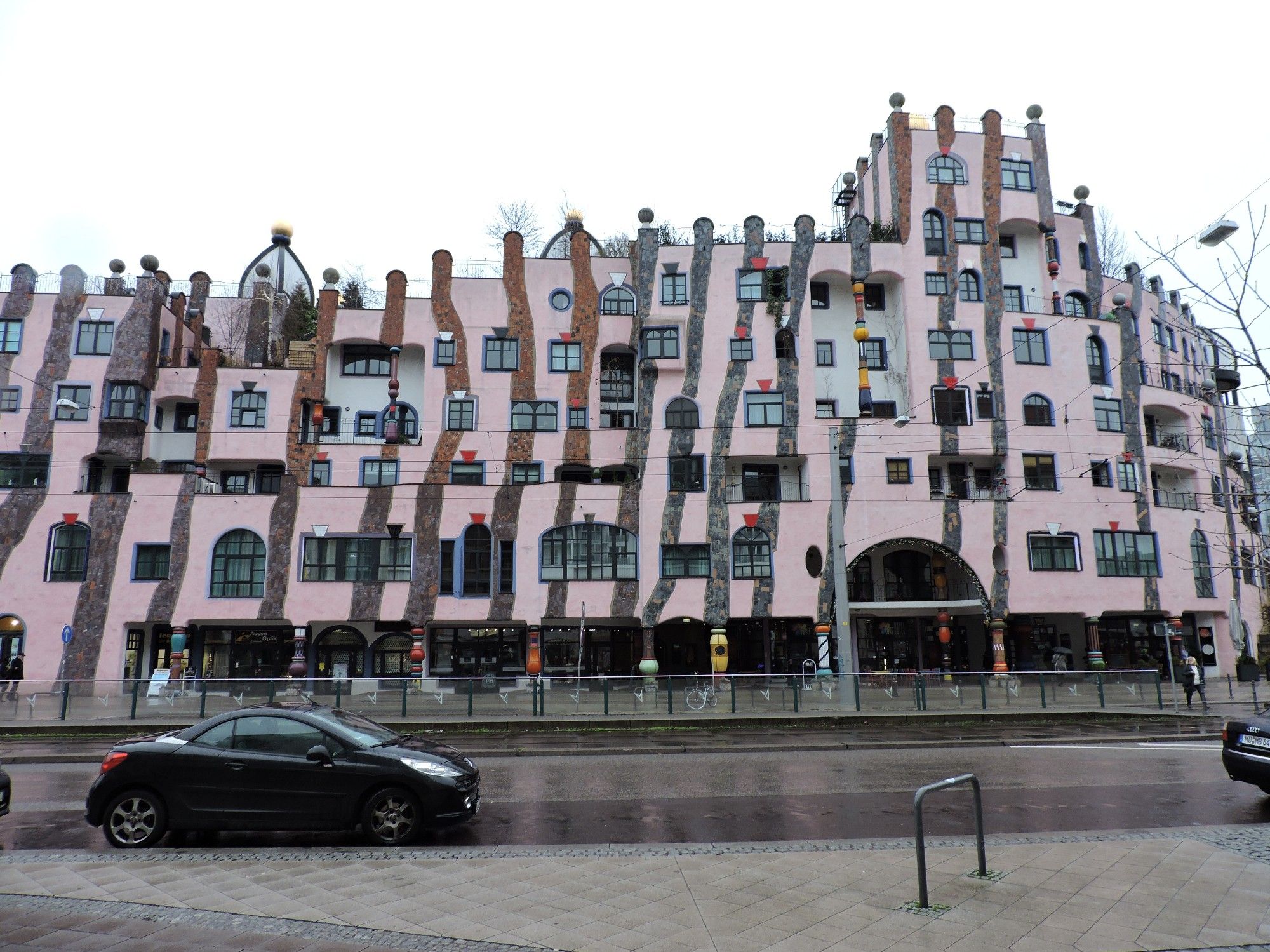 Eine verspielte hellrosa gestrichene Fassade eines Gebäudes in Magdeburg. Die Straße davor ist nass und ein schwarzes Auto, so eine Art Sportwagen parkt im linken Bildbereich.