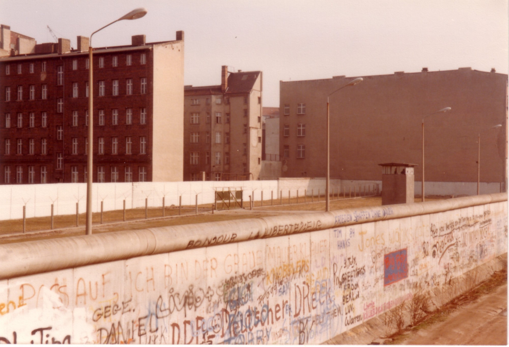 Berliner Mauer. Dahinter Todesstreifen mit großen Laternen und im Hintergrund Wohnhäuser 