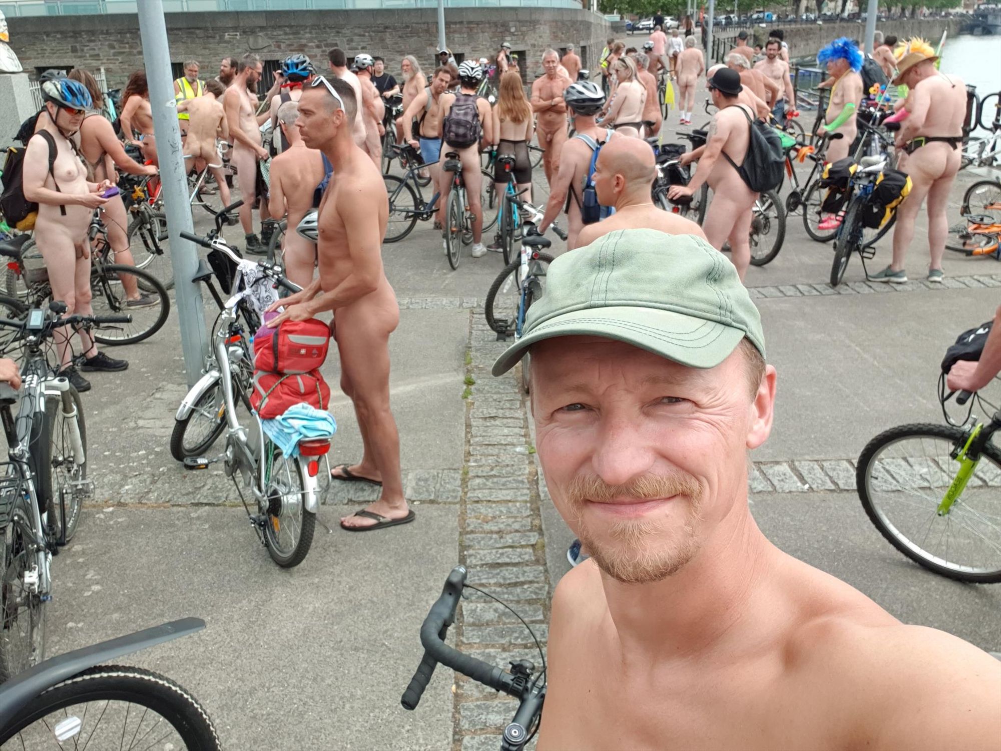 Selfie of a white man wearing a green cap. Behind him are other people with bikes, mostly naked.