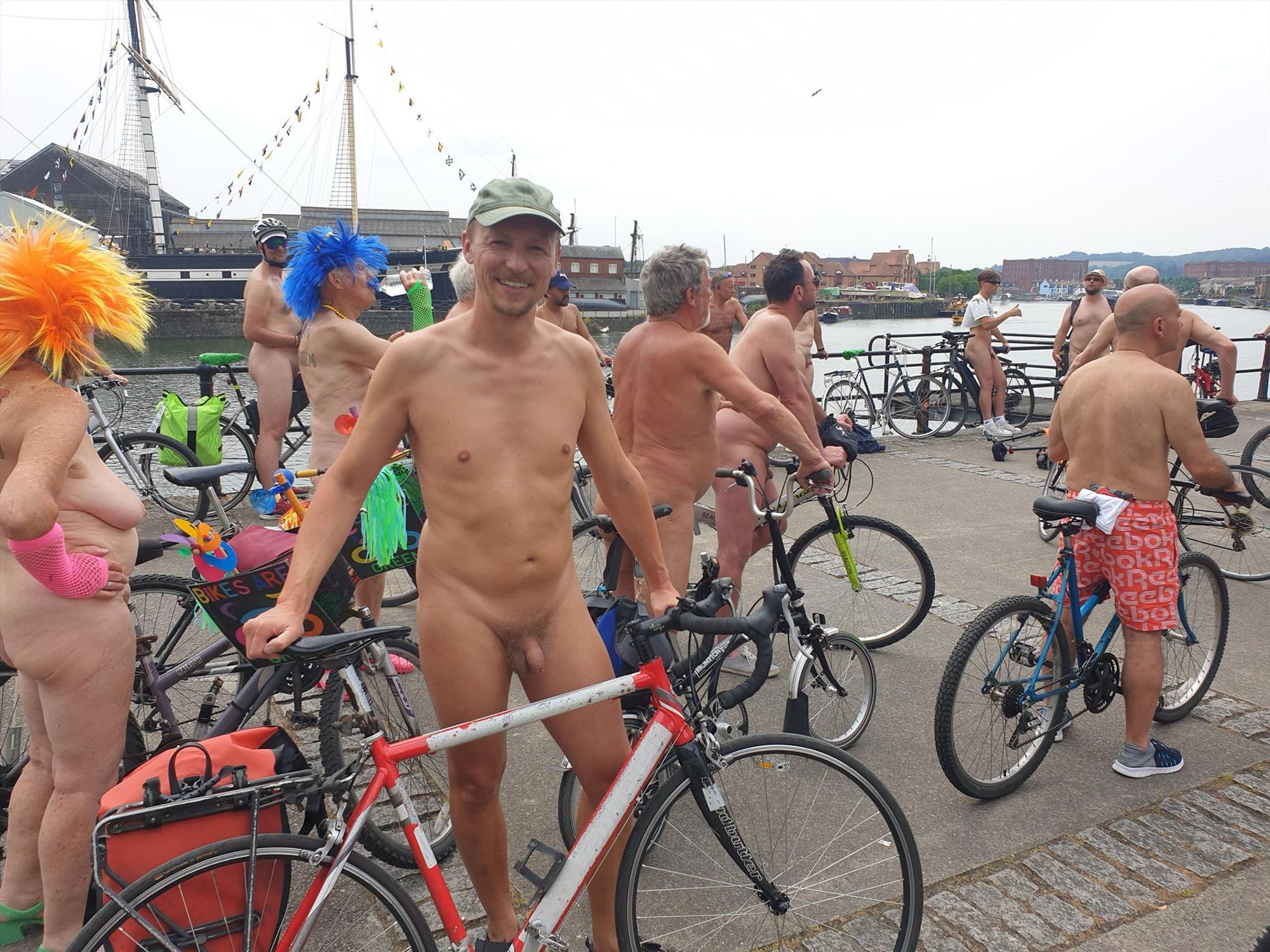 Photo of a naked white man stood beside a bike. Around him there are other people with bikes, mostly naked. They're all on a path next to a waterway - it's Bristol's harbourside. The Victorian steamship SS Great Britain is visible in dry dock in the distance.