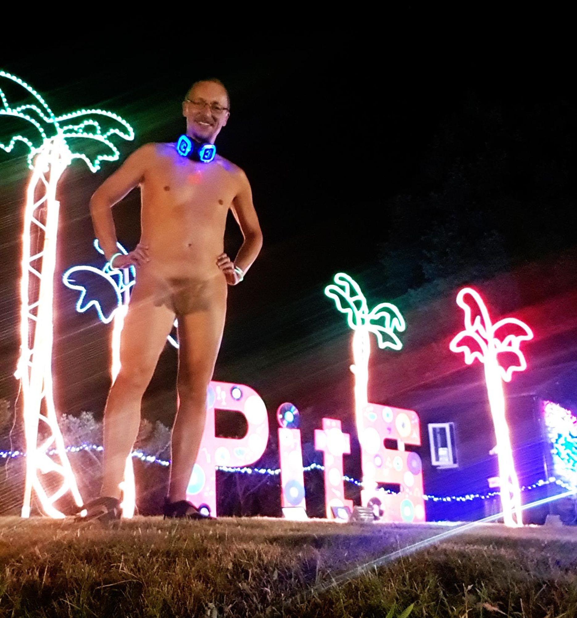 Photo of white man stood naked in a festival field at night. He has his hands on his hips, and glowing headphones are resting around his neck. Behind him, there are illuminated palm trees and a sign saying PitS.