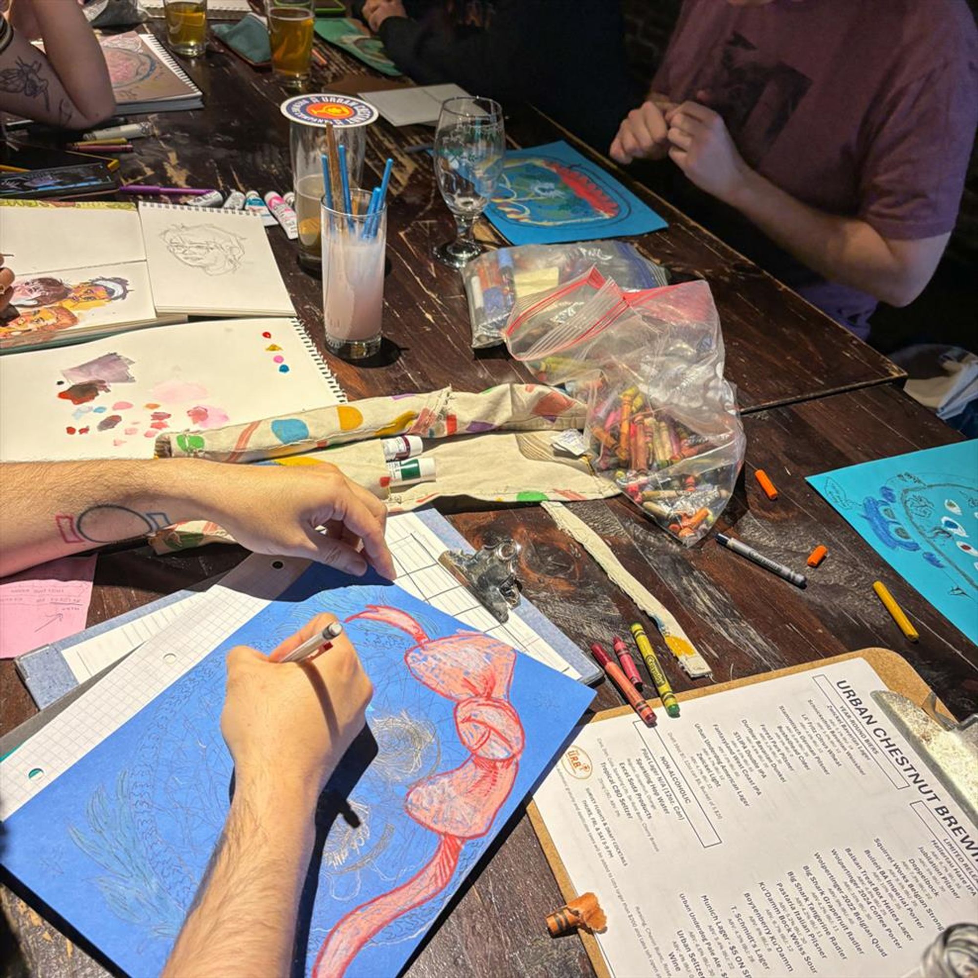 Close up of hands working on drawings with crayons, paint, and other media.