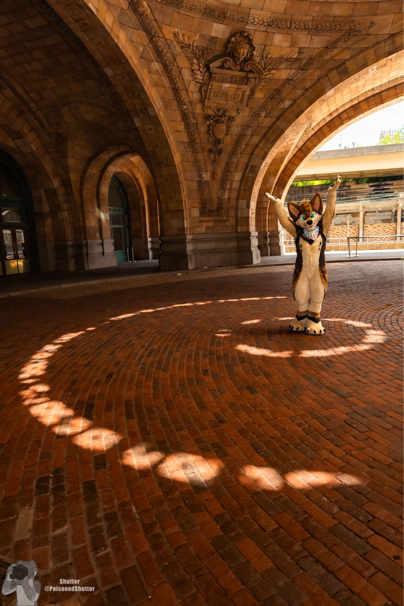 A coyote fursuiter stands in the rotunda of a historic train station. The glass peak of the rotunda is allowing arcs of sunlight to shine through that surround the subject